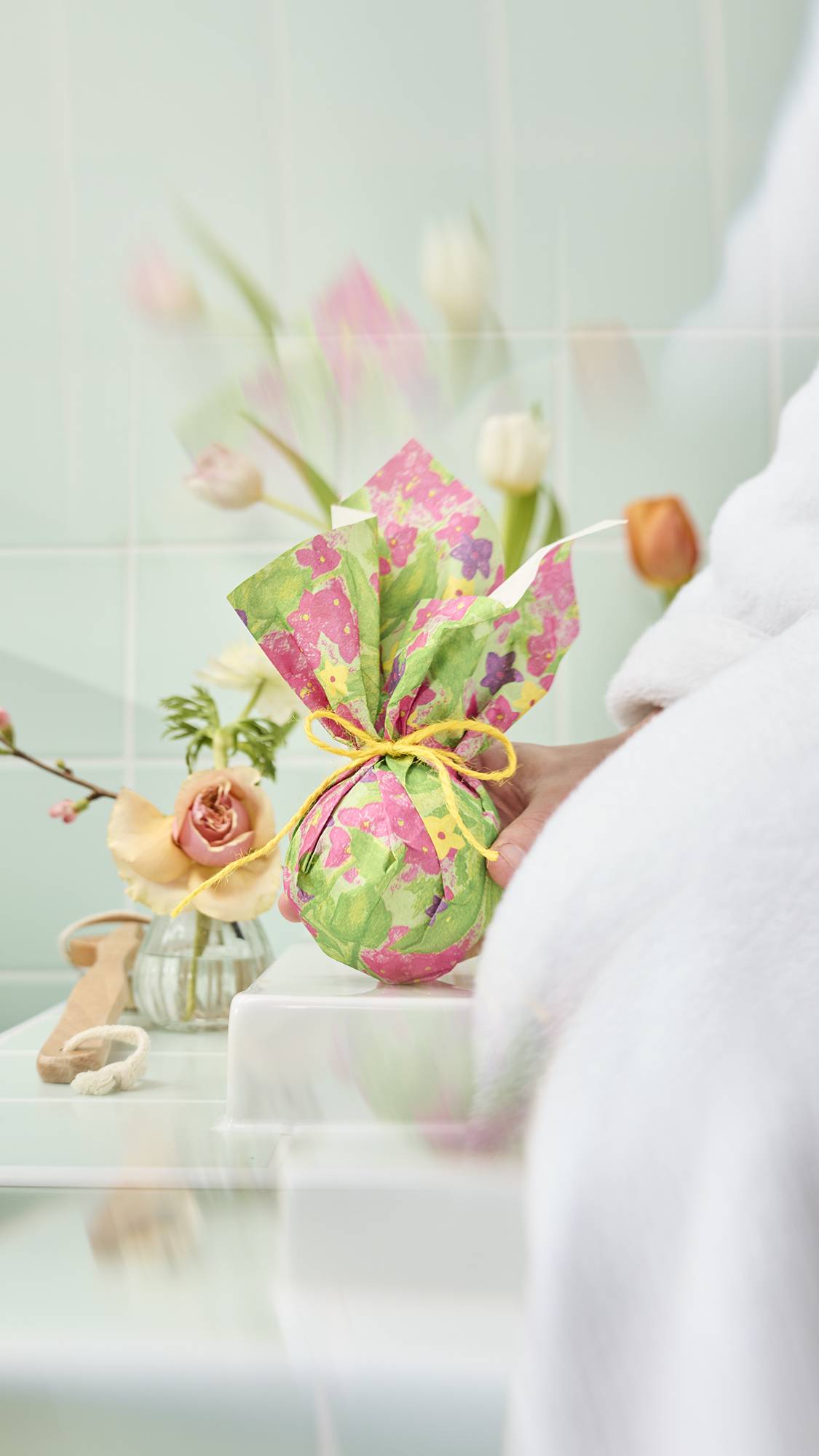 The image shows a round bath bomb wrapped in a sheet of "Garden in Spring" Lokta paper. The wrap is secured at the top with a yellow twine bow, creating a decorative gathered effect. The background shows a soft, botanical-themed bathroom setting.