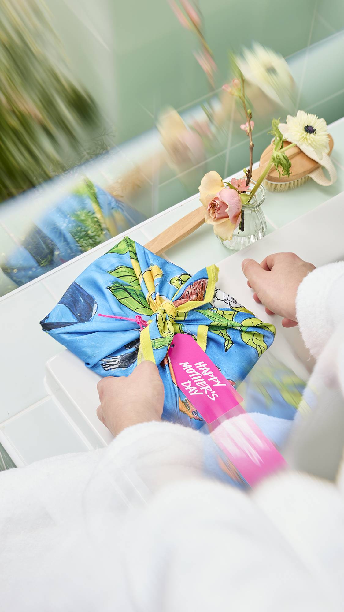 A model in a white dressing gown ties a blue fabric "Garden Mural" knot-wrap, featuring a whimsical nature design, over a gift. A pink "Happy Mother's Day" tag is attached to the knot. The scene is set in a bright bathroom with soft floral accents.