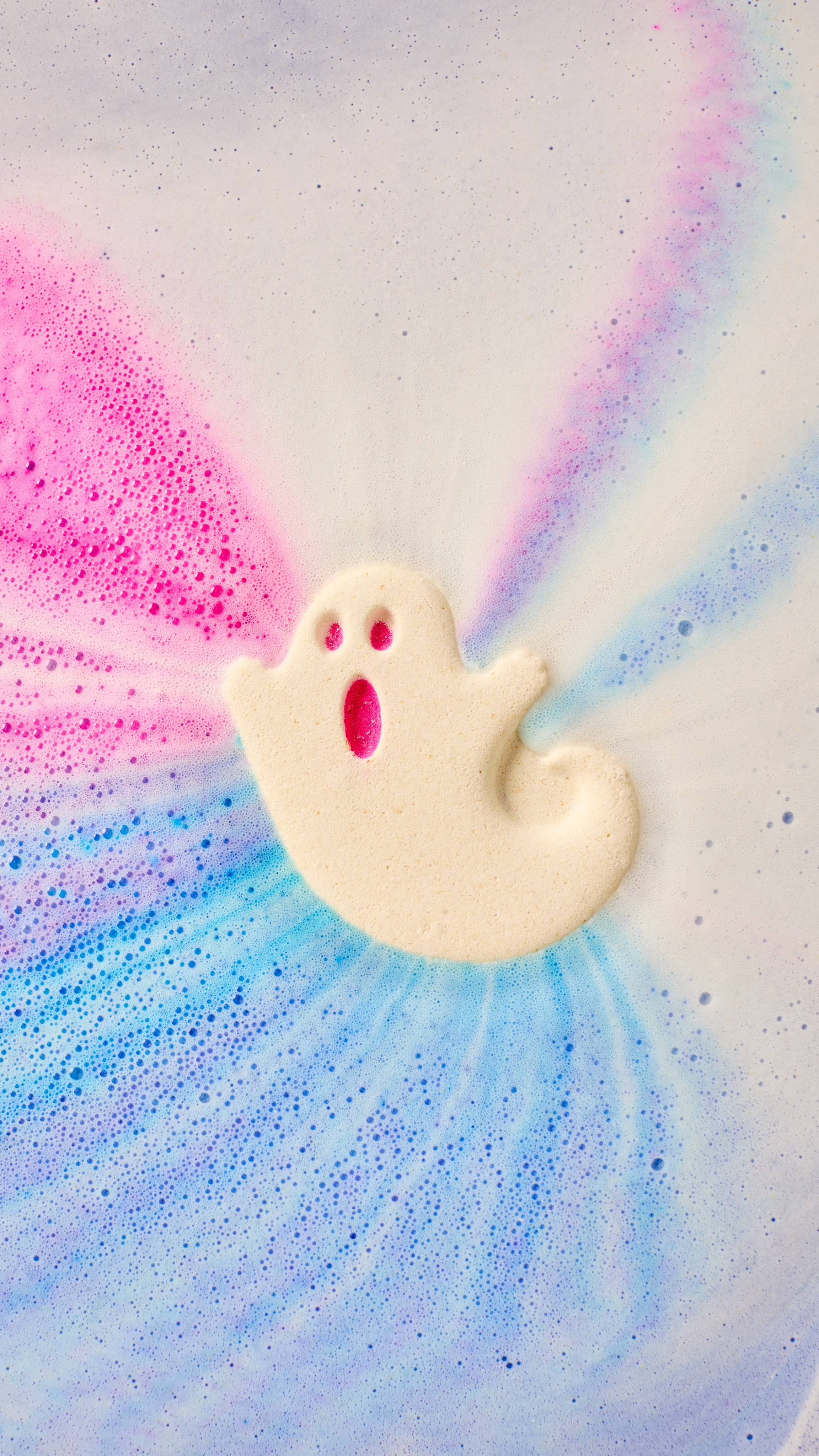 The image shows a white 'Ghostie' bath bomb, with pink eyes and mouth, dissolving in swirling bath water. Vibrant pink and bright blue hues blend with white foam, forming dynamic, concentric bath art patterns.