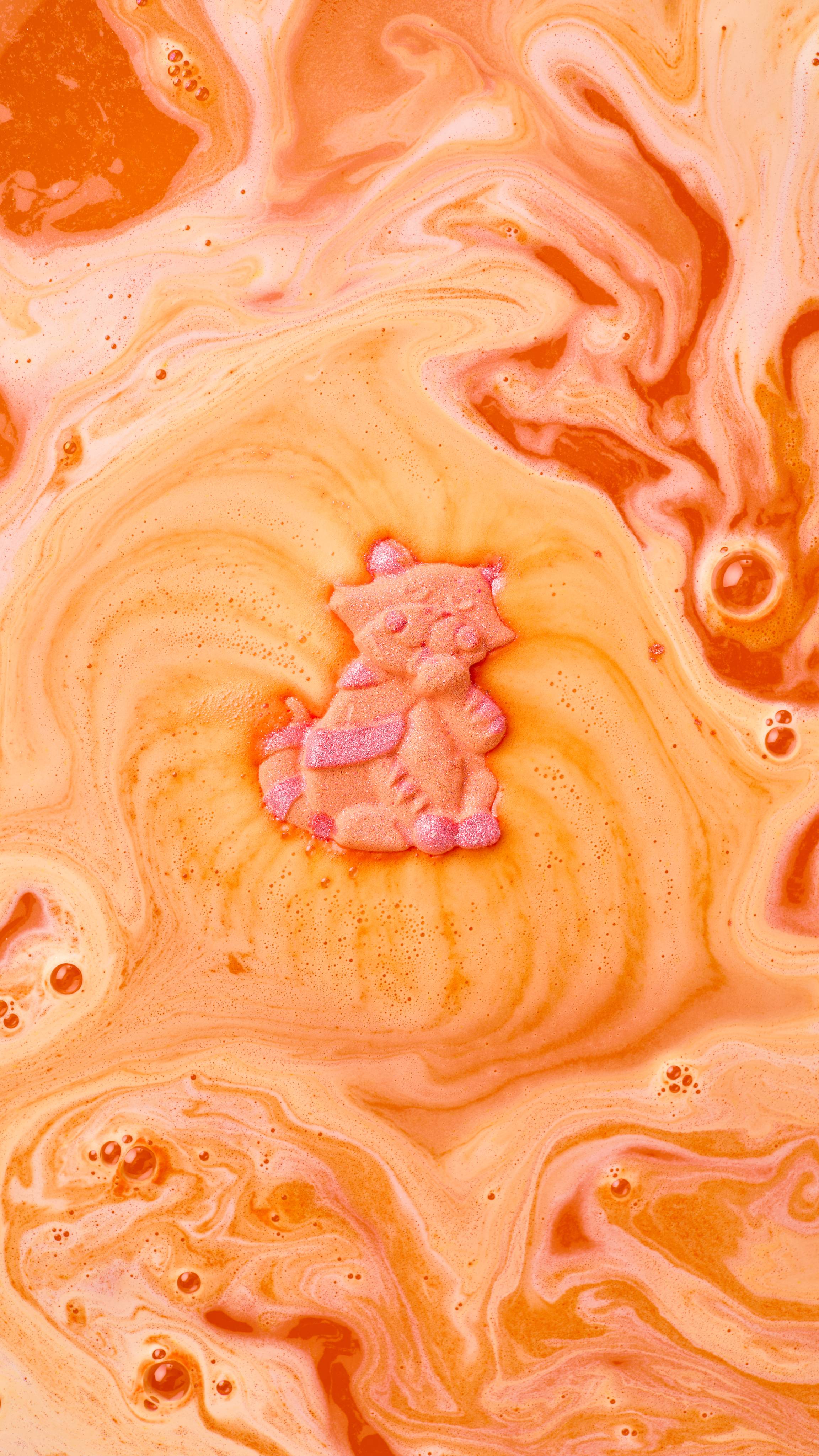 The image shows a vibrant orange 'Ginger Tom' bath bomb, with pink markings, fizzing in swirling yellow and orange bath water. The water forms a vibrant spiral around the dissolving cat, blending with light foam and bubbles.