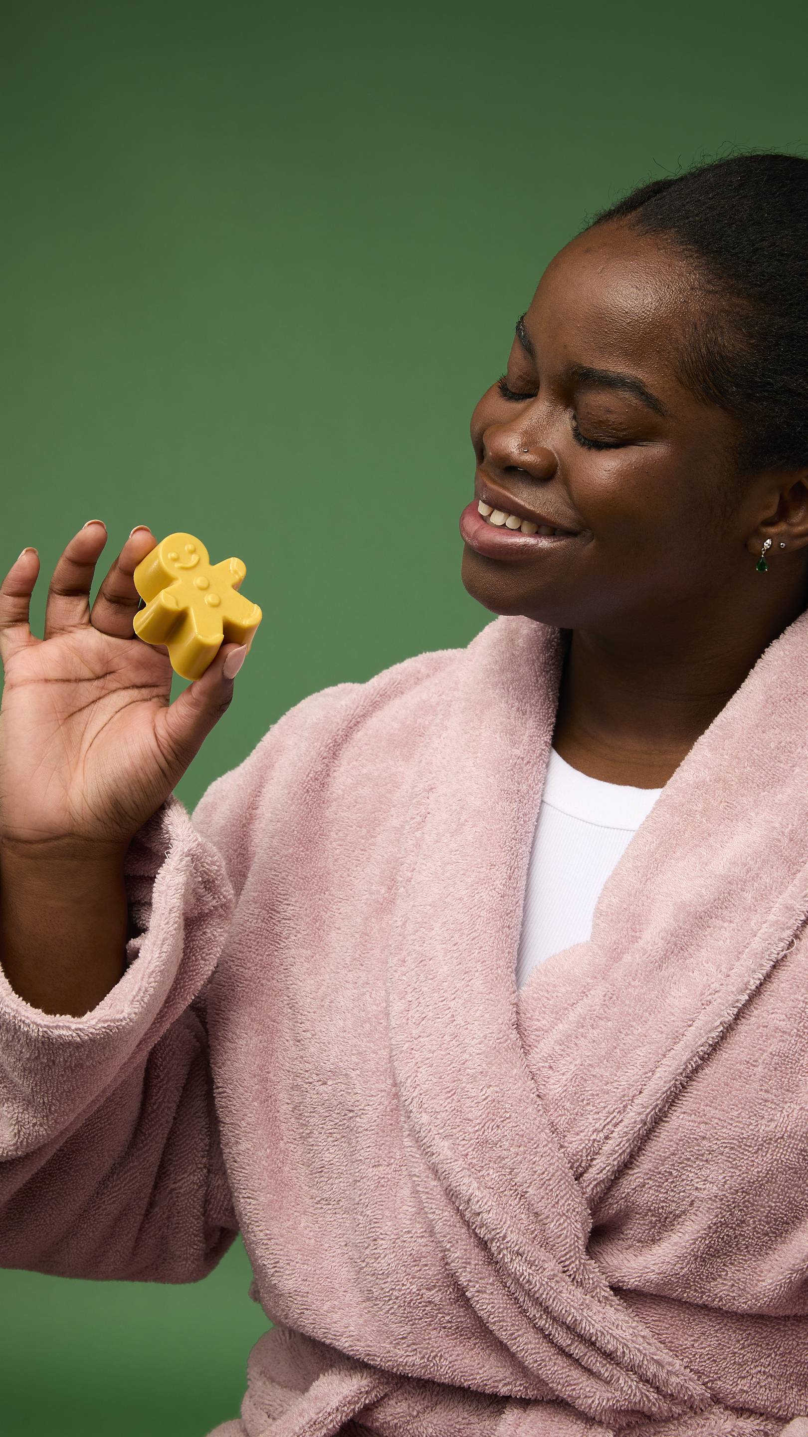 The image shows a model with dark skin, wearing a pink bathrobe, holding a small, golden-yellow Gingerbread Man gummy facial wash. They hold the product up, with their eyes closed, appearing to smell it. The background is solid green.