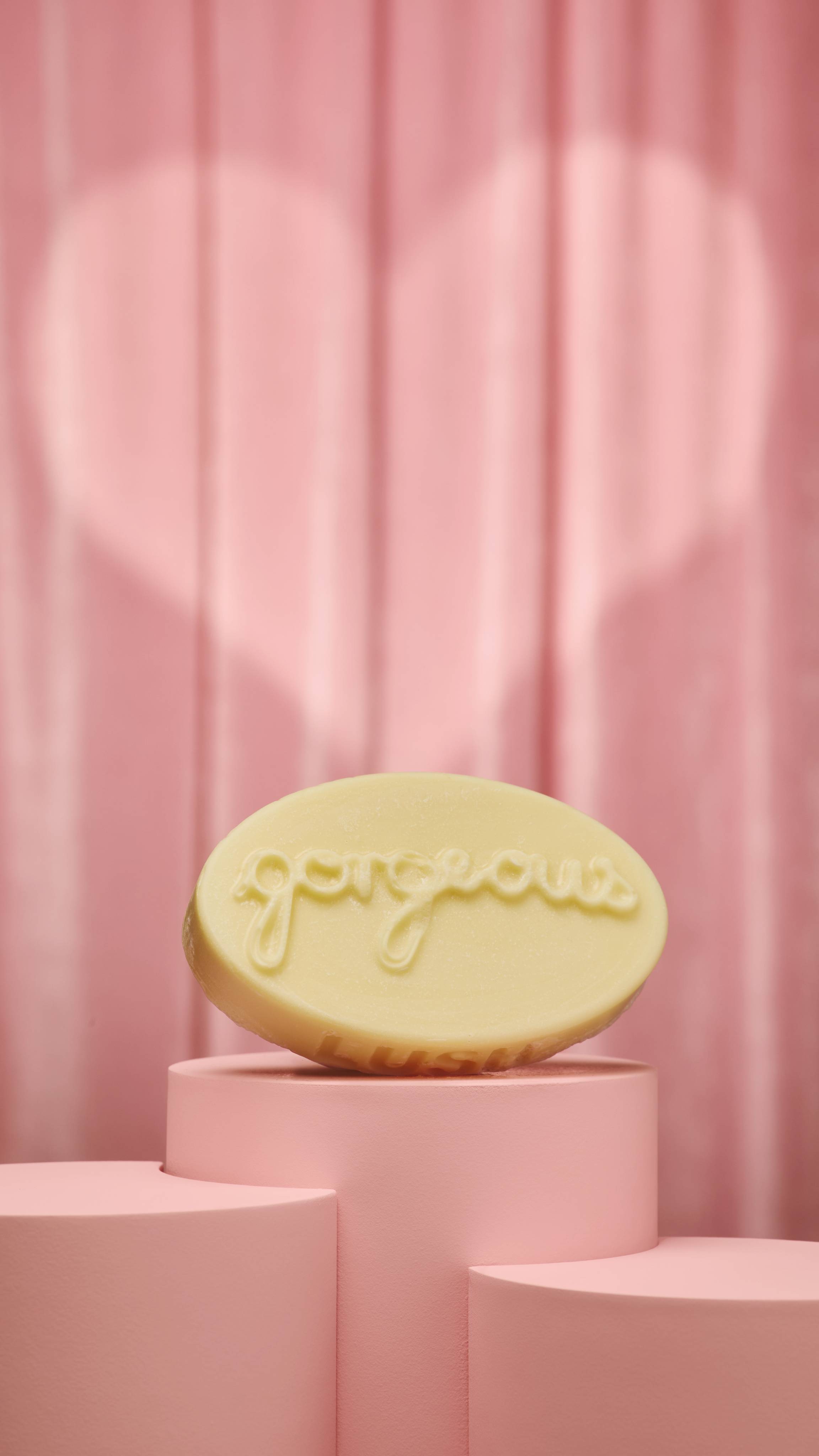 A cream-coloured, oval massage bar with the word "gorgeous" embossed on its top surface sits on a pink, cylindrical podium. The background consists of soft pink curtains with a large, subtle heart-shaped spotlight illuminating the product.