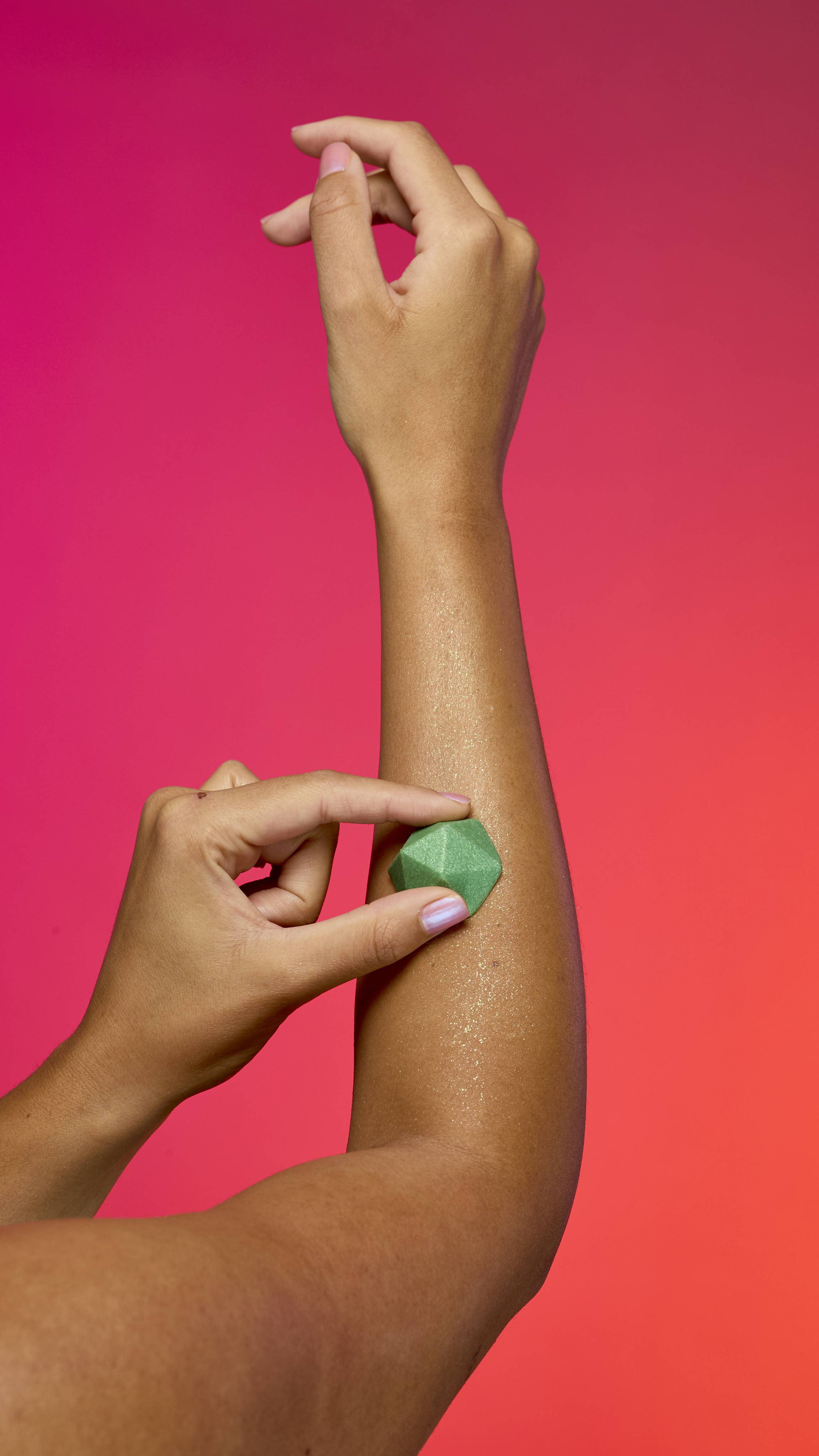 Two hands with tan skin apply the faceted, pyramid-shaped Green Glitterbug body bar to an arm. The skin has a sheen of plastic free glitter against a vibrant gradient of magenta, pink, and orange.