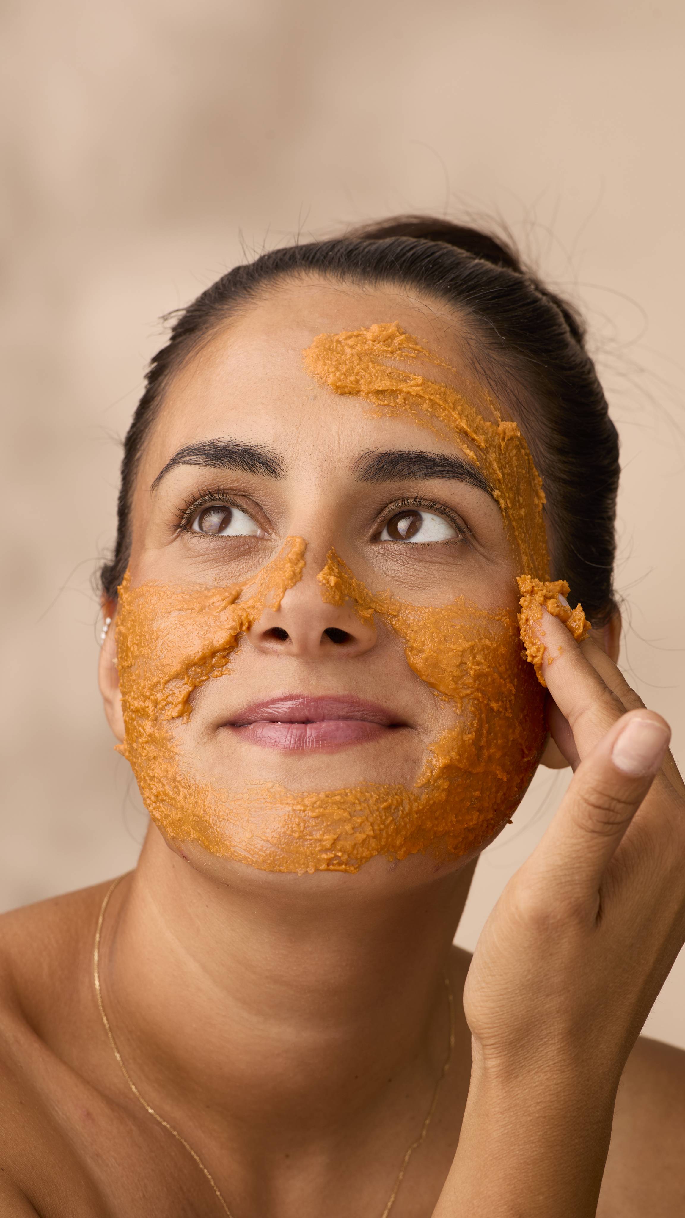 A woman with dark hair tied back looks up and smiles gently while applying a thick layer of bright orange Lush Halloween Queen face mask. The textured paste covers her jaw, nose and cheeks as her fingers smooth it across her forehead