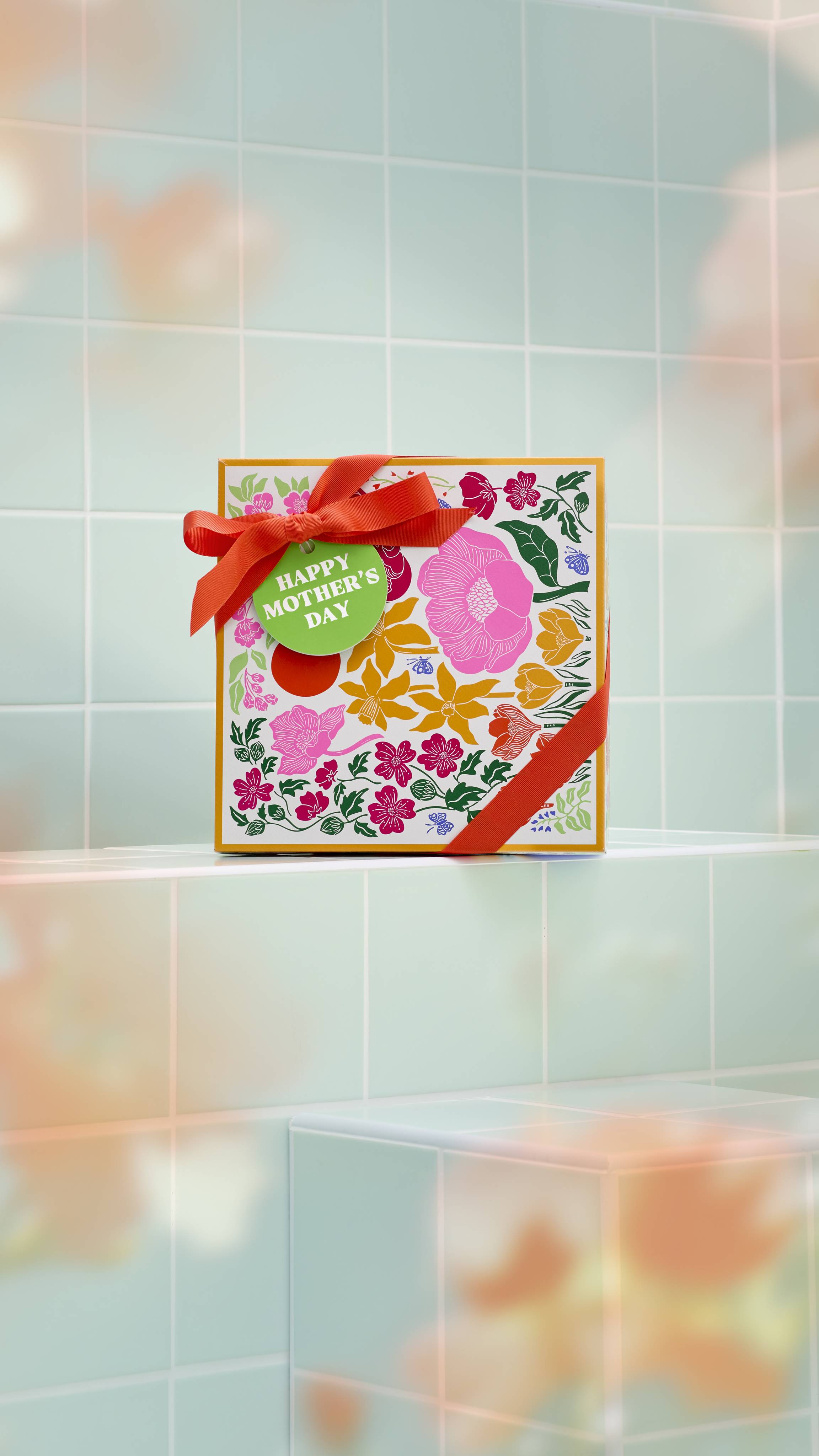 A square Lush "Happy Mother’s Day" gift box with a colourful floral design and red ribbon stands on a pale green tiled ledge. Soft, hazy floral shadows frame the scene.