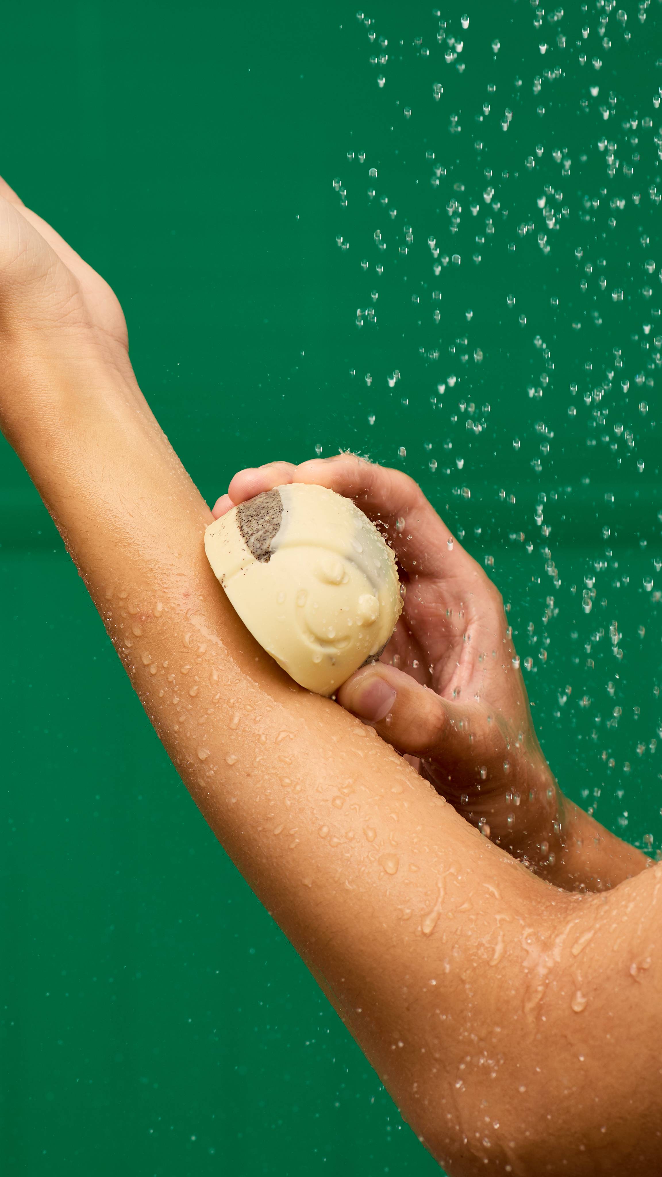A hand is using a bee-shaped LUSH Humbug Scrubee solid body scrub on a wet forearm. The pale yellow scrub has a dark exfoliating stripe on the back. Water droplets are visible on the skin and in the air against a vibrant green background.