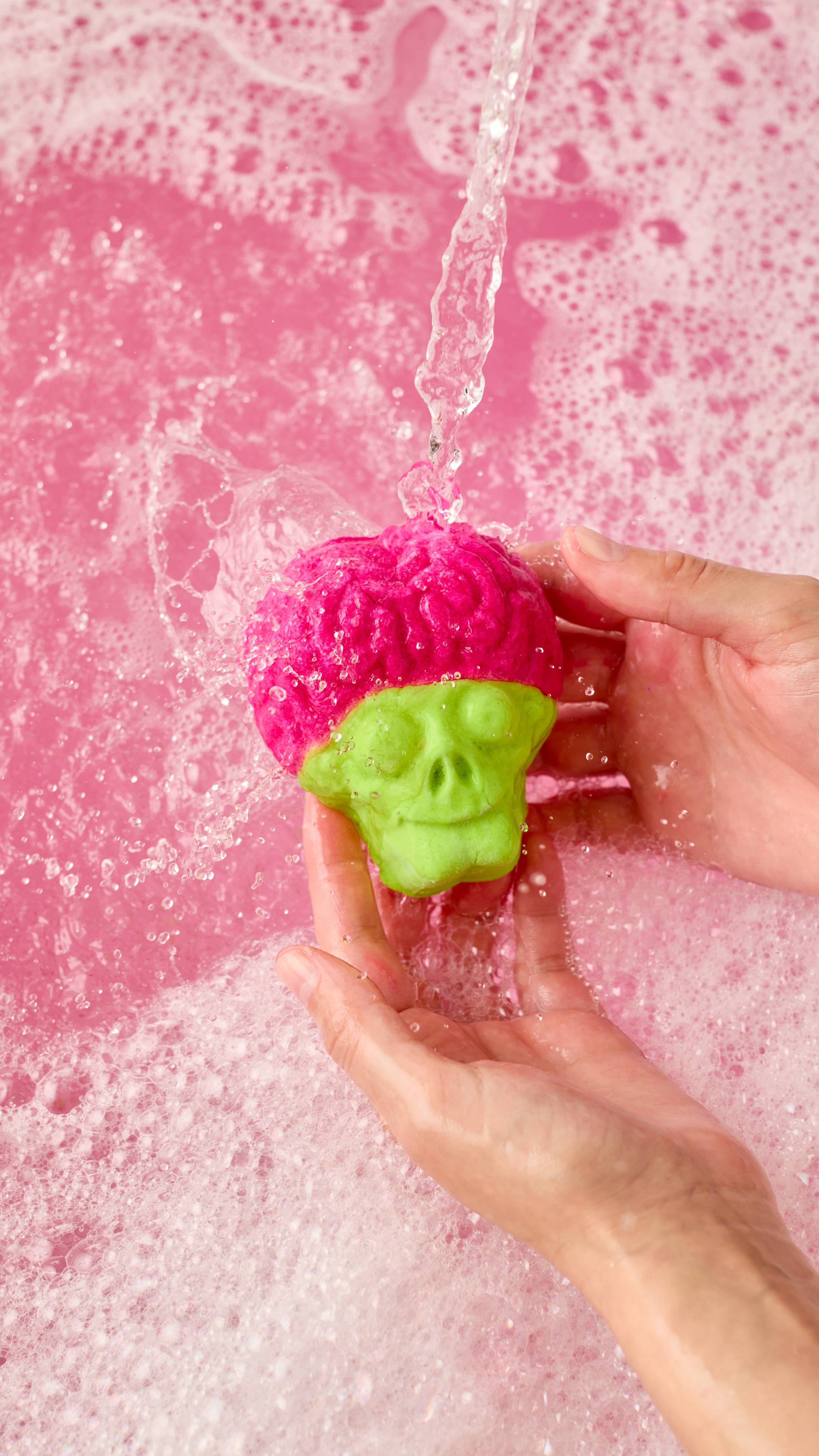 The image shows two light-toned hands holding a pink and green 'Invaders from Mars' bubble bar. Water pours onto the bar, generating vibrant pink bath water with bubbles and thick foam.