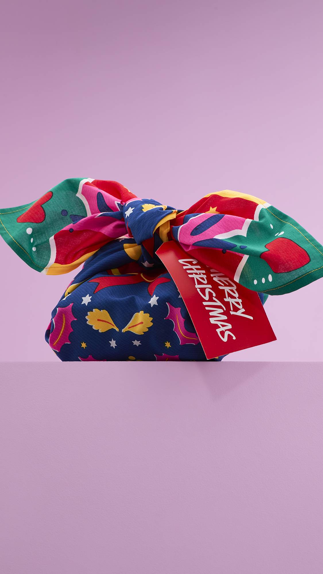 A vibrant gift wrapped in a Lush "Christmas Deer" Knot Wrap. The reusable fabric features a festive pattern of a deer, stars, and holly. It is tied with a knot and has a red "Merry Christmas" tag.