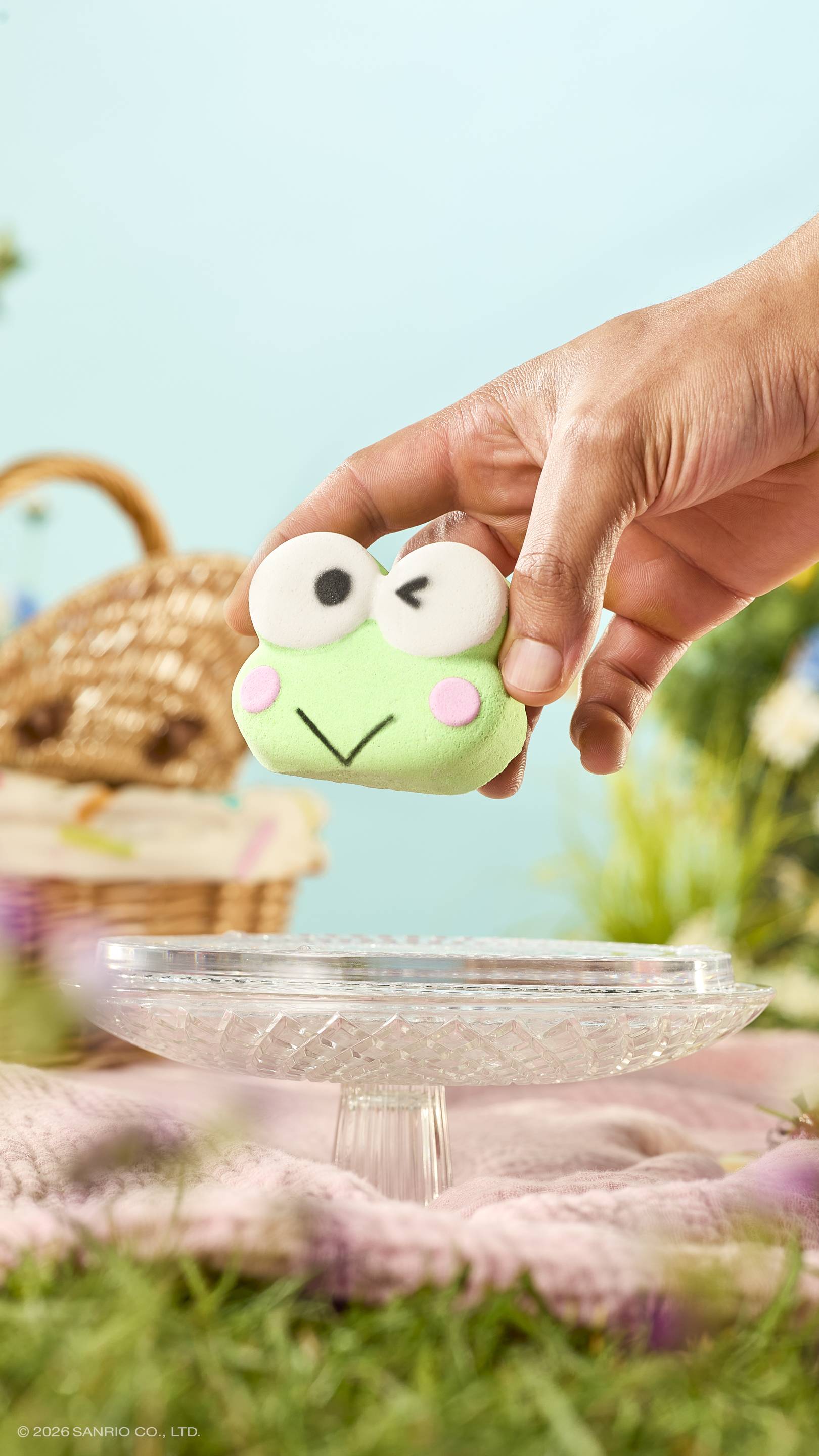 A hand holds a green, winking frog-shaped bath bomb over a glass cake stand. The scene is a sunny outdoor picnic with a wicker basket, pink blanket, and grass against a soft blue sky.