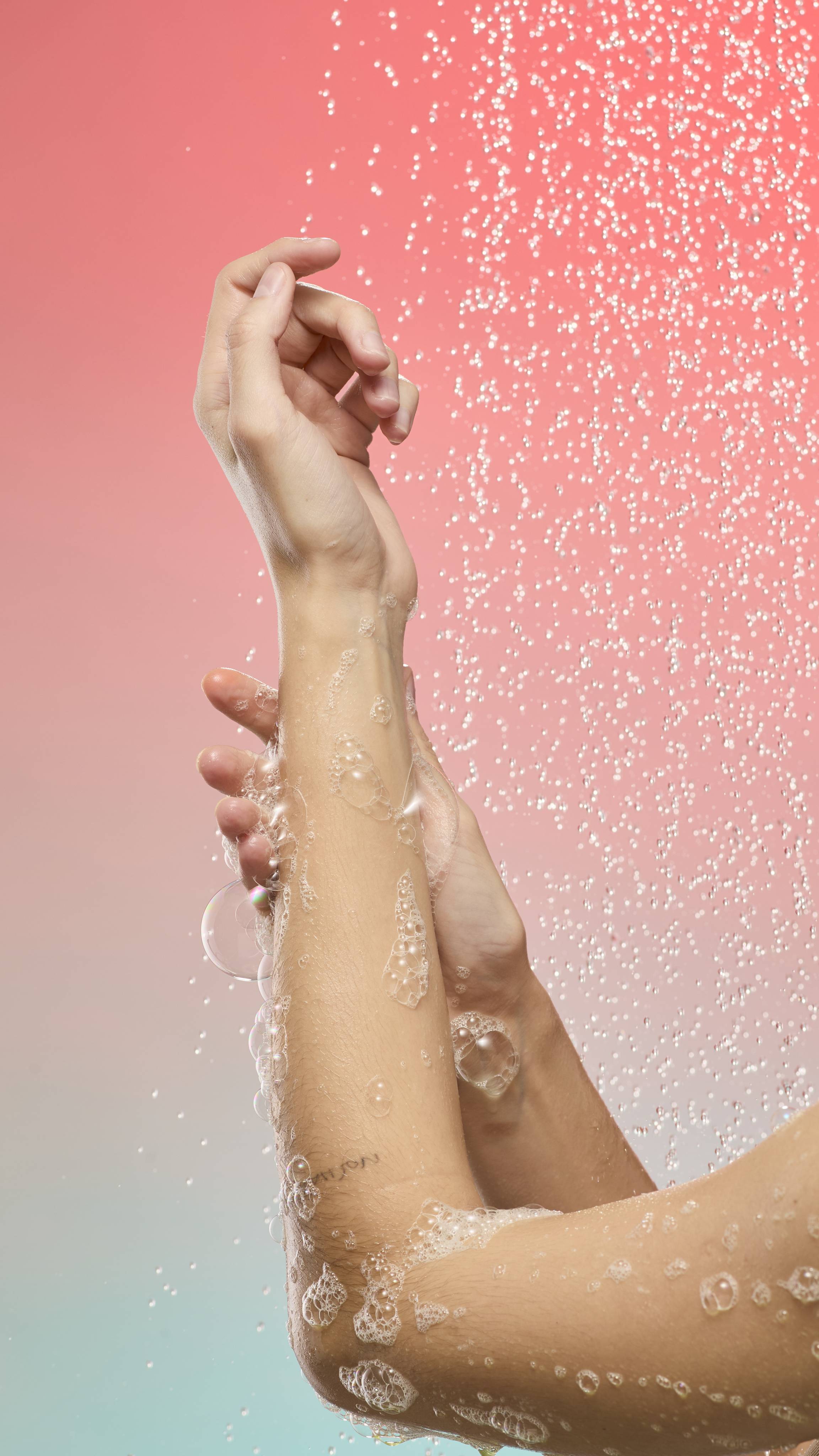 Two arms covered in foamy soap bubbles from Lemon Sherbet shower gel are held under a spray of falling water droplets. The background is a soft peach-to-blue gradient.