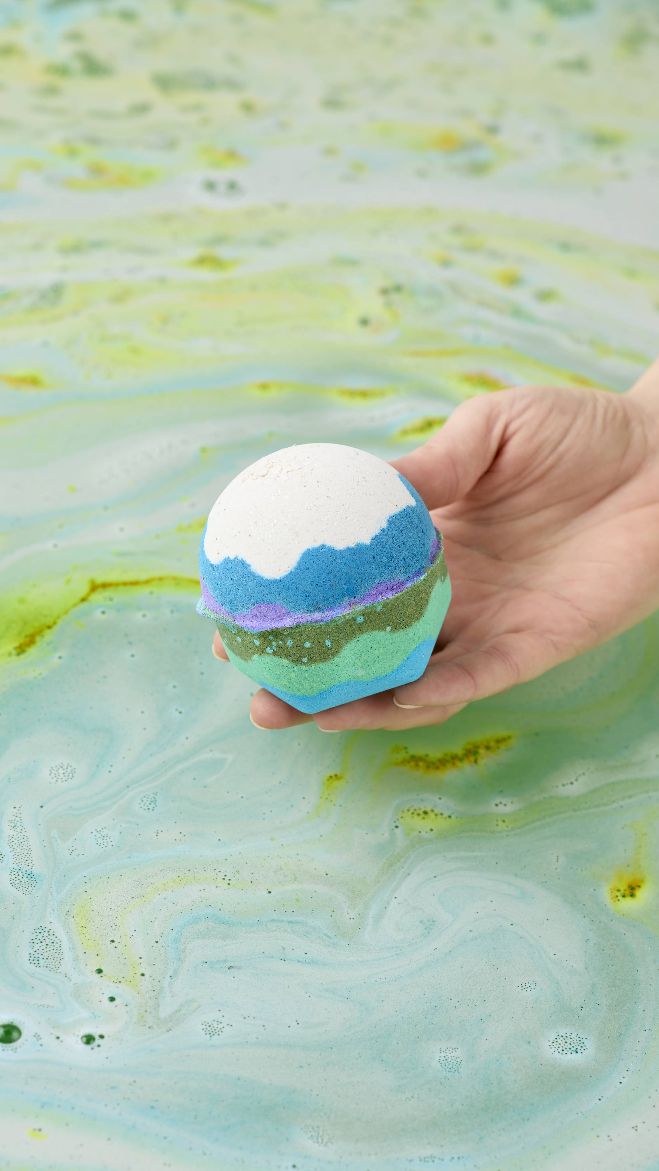 A hand holds the "Lola's Aurora Borealis" bath bomb, with layers of white, blue, purple, and green over swirling light green and turquoise water in a tub. The water is covered in fine, frothy white bubbles and marbled with streaks of yellow.