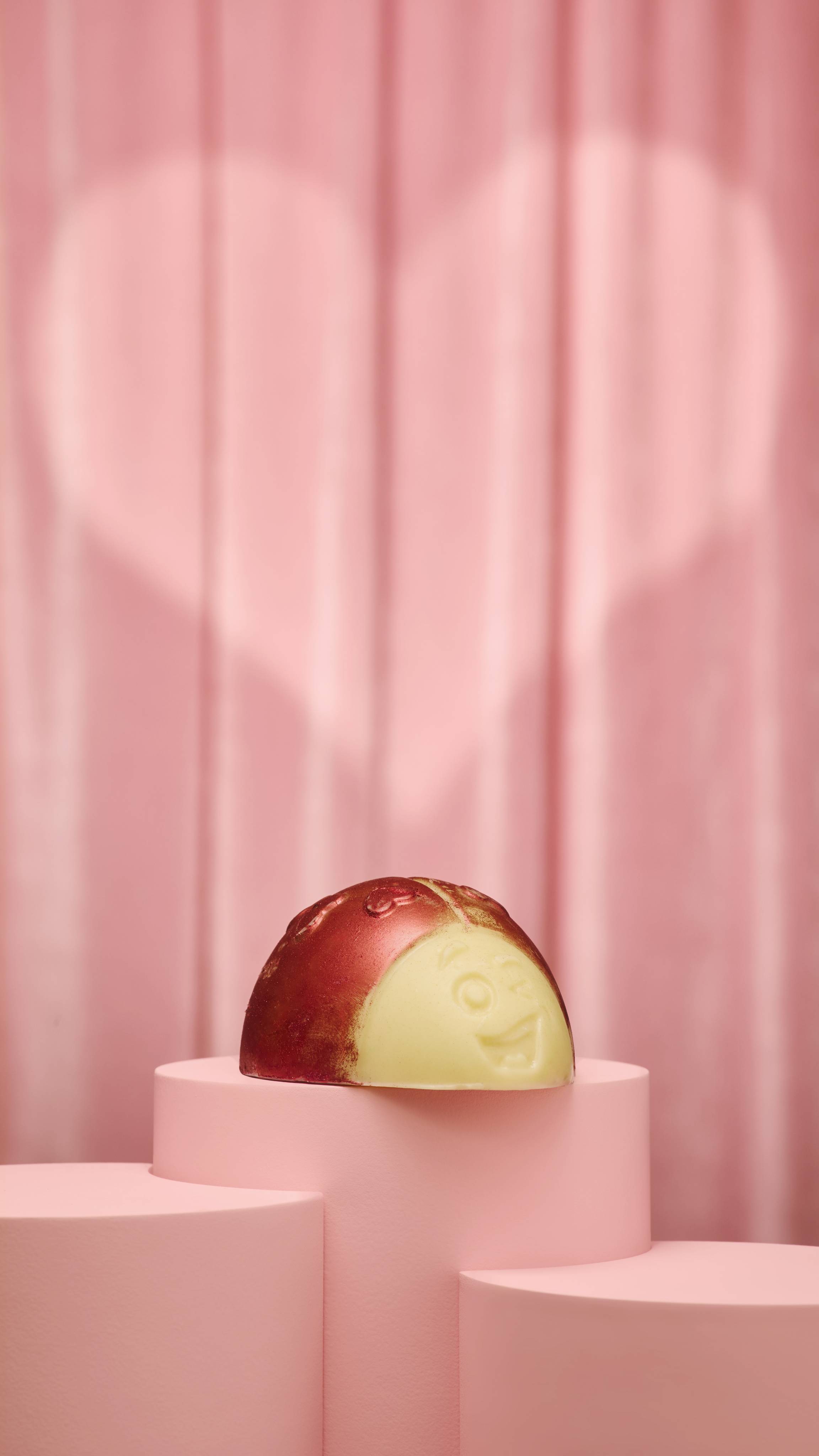 The image shows the large, creamy, ladybug-shaped Love Bug Scrubee solid body scrub. The product features heart-shaped spots and red lustre while sitting on a pink podium on a pink fabric background and a heart-shaped spotlight. 