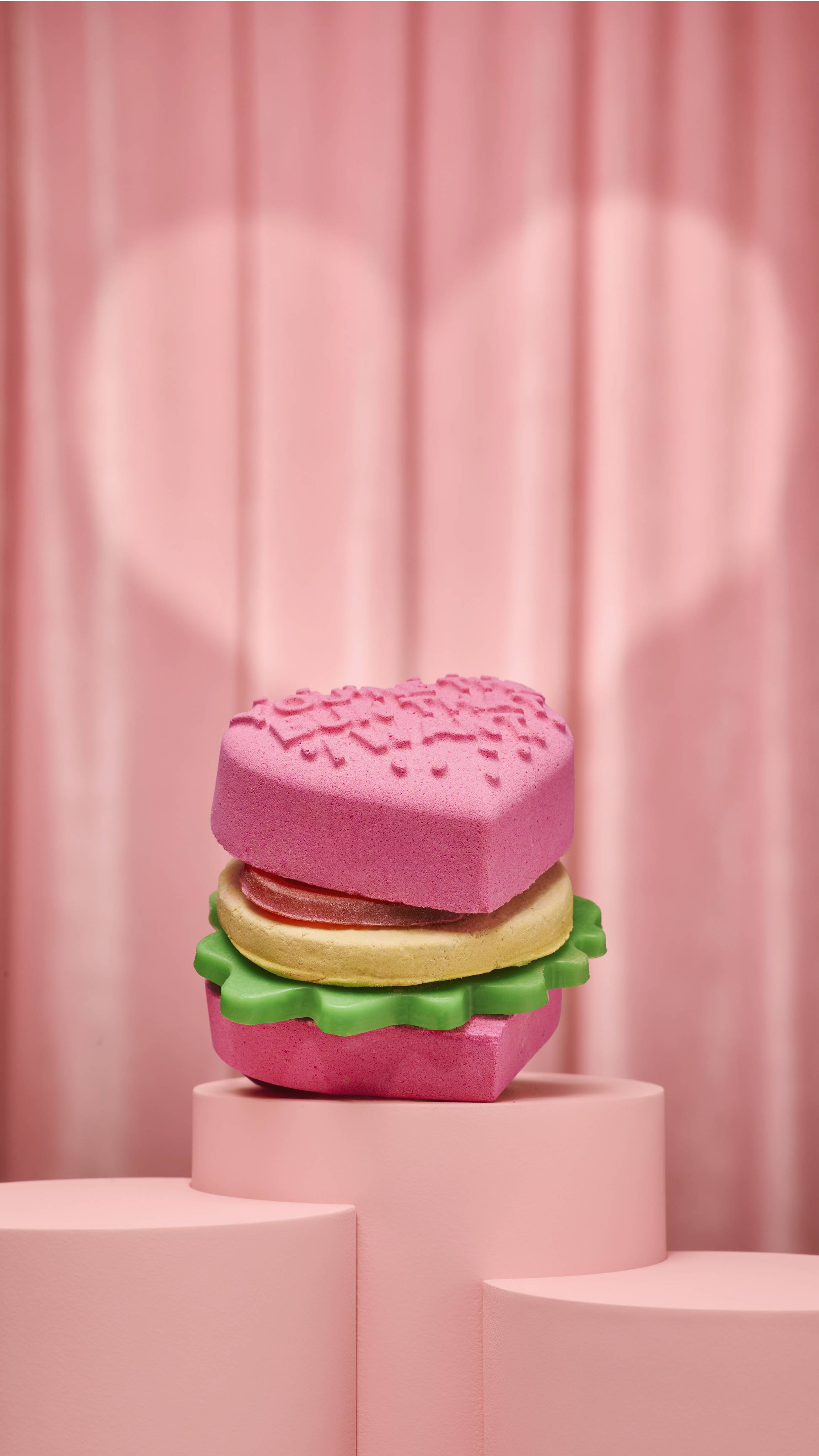 A heart-shaped Love Burger bath bomb sits on a pink podium. It consists of pink heart buns, a yellow patty bubble bar, a red tomato-shaped soap layer, and a scalloped green lettuce soap layer.