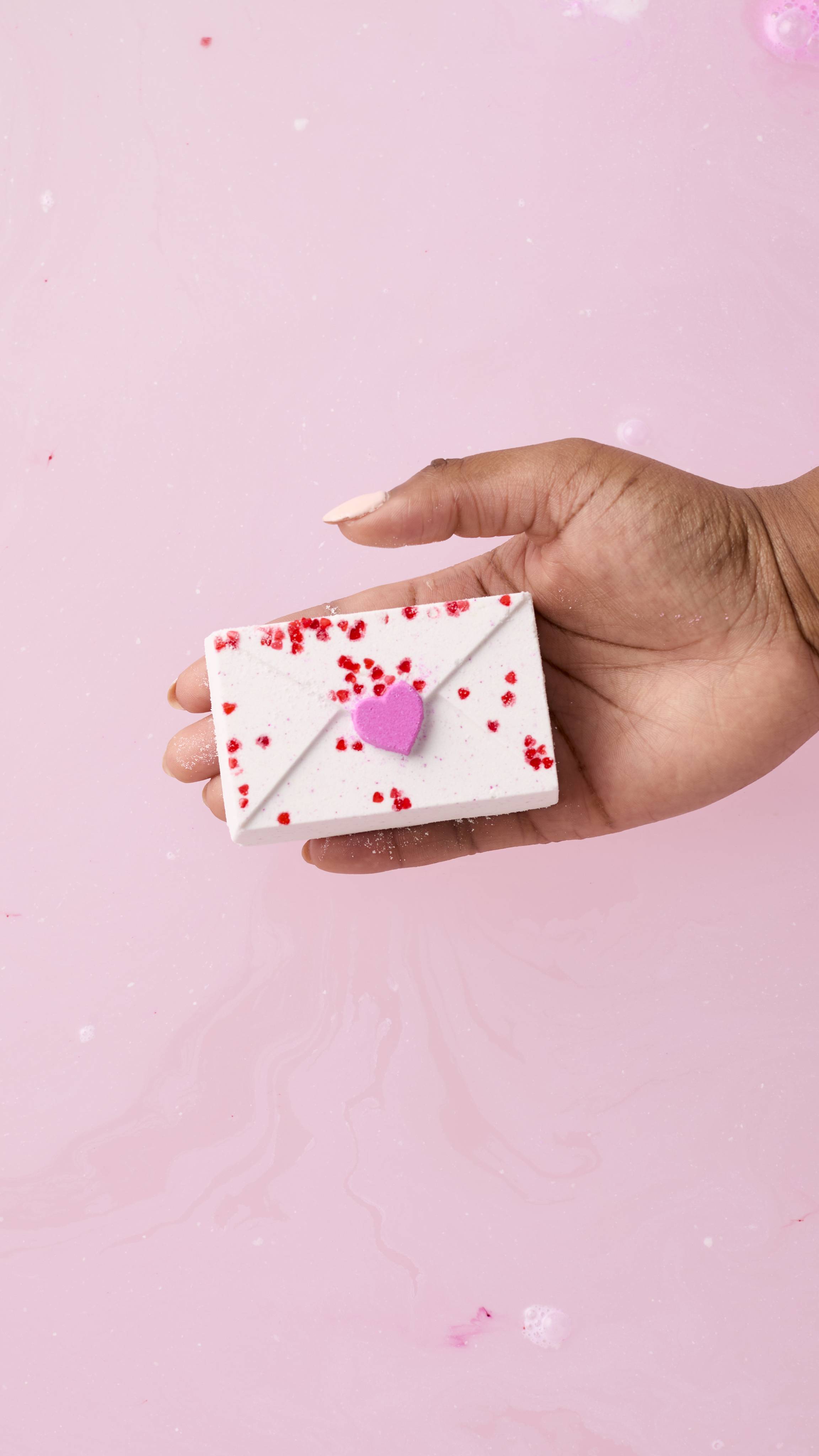 A person’s hand holds a white, envelope-shaped bath bomb over soft pink water. The bath bomb is decorated with tiny red flecks and a single raised purple heart, designed to look like a sealed love letter.