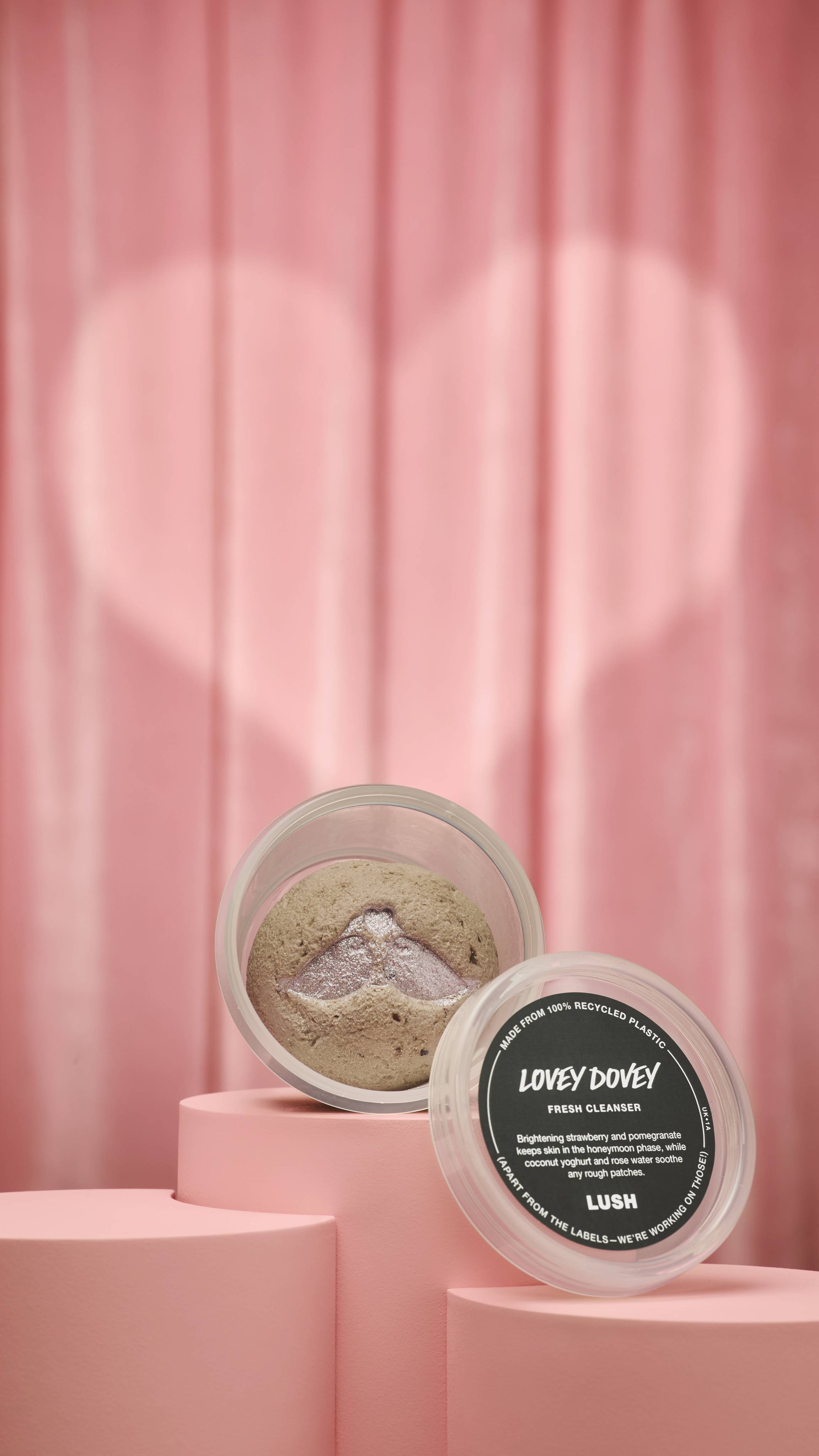 The open pot of Lovey Dovey Fresh Cleanser sits on a pink pedestal, displaying the textured product inside and the black lid with white text against a soft pink curtain background illuminated by a heart-shaped spotlight.