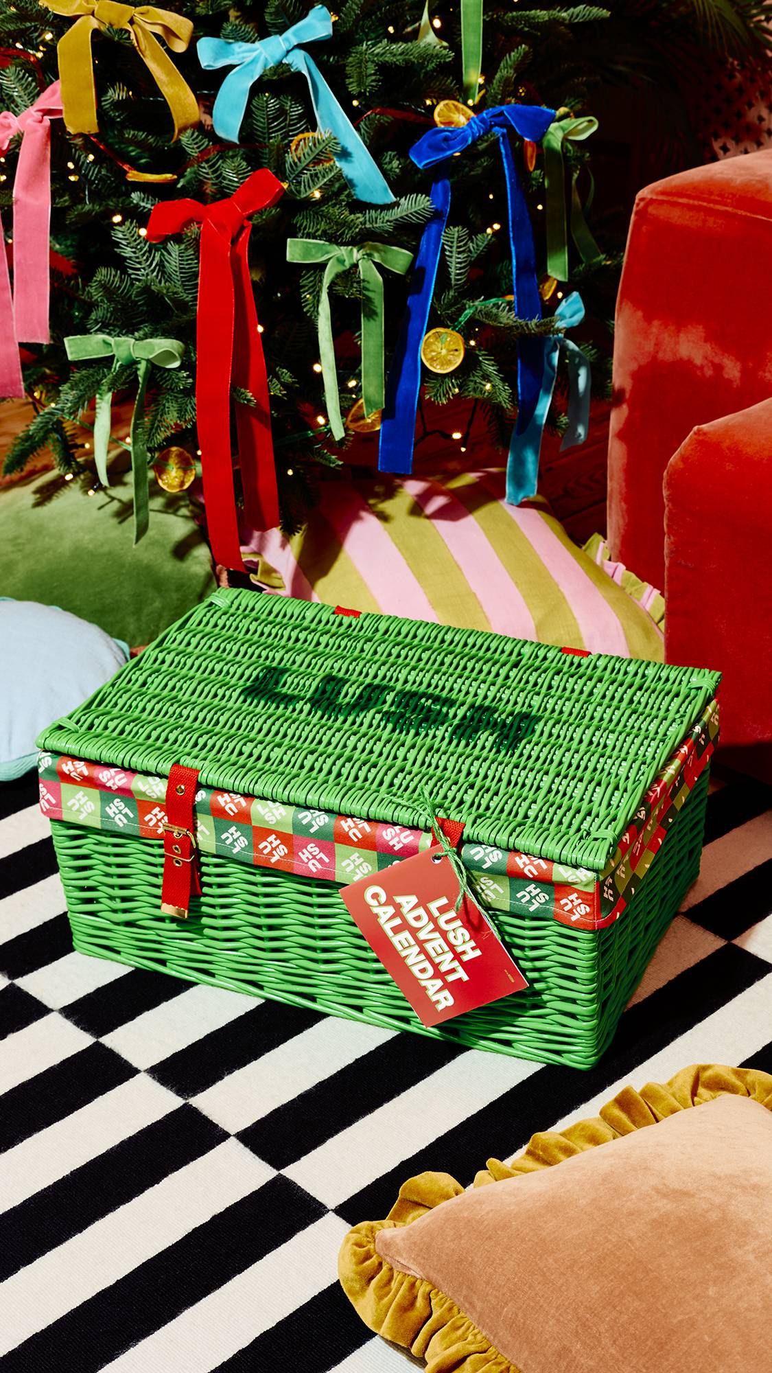 A closed green wicker hamper with red straps and a red tag reading "Lush Advent Calendar" sits on a black-and-white striped rug in front of a Christmas tree adorned with colourful ribbon bows.