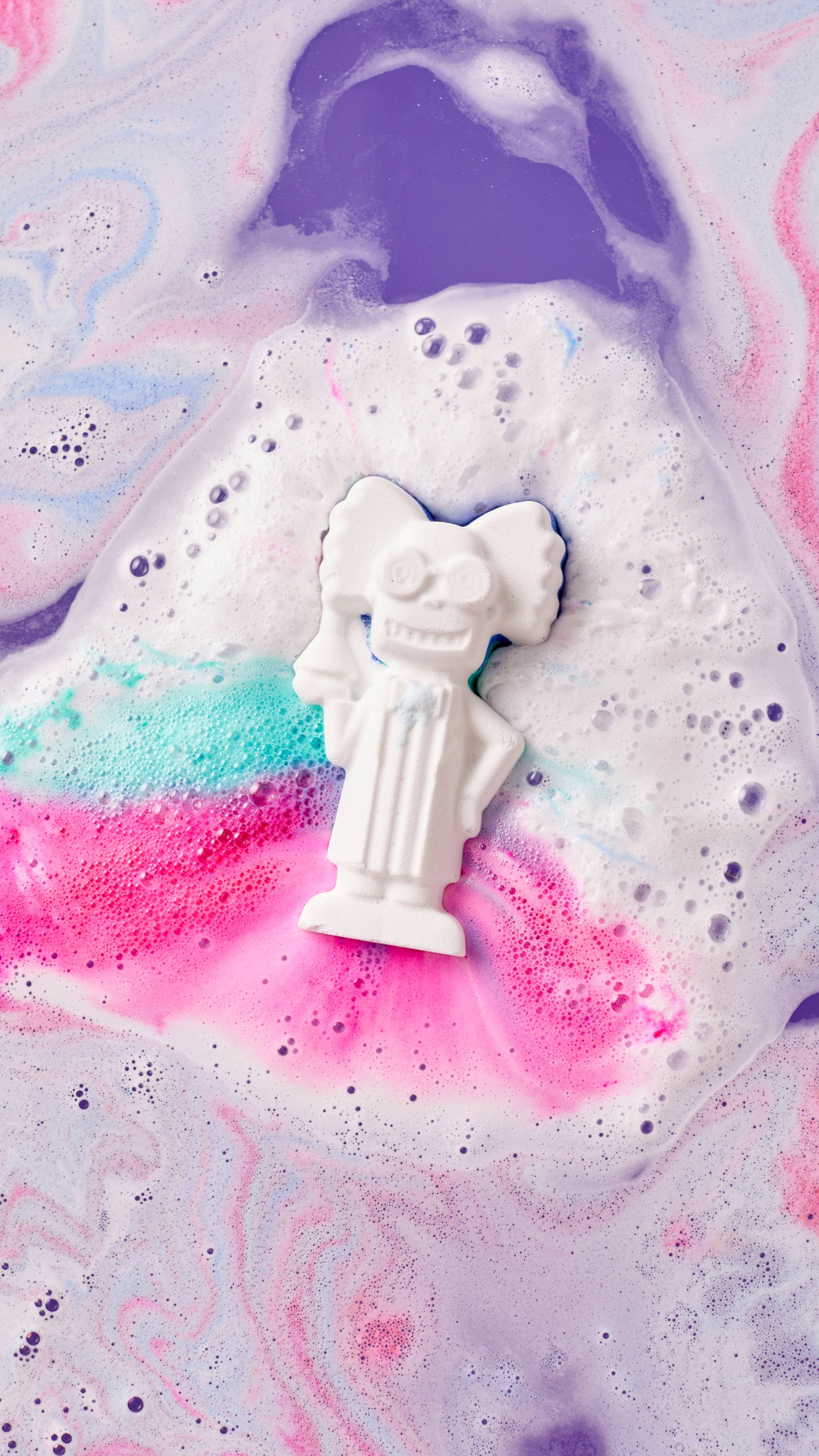 The image shows a white 'Mad Scientist' bath bomb, with blue and pink streaks, fizzing in vibrant, multi-coloured bath water. Pink and turquoise foam surrounds the character, blending with purple hues.
