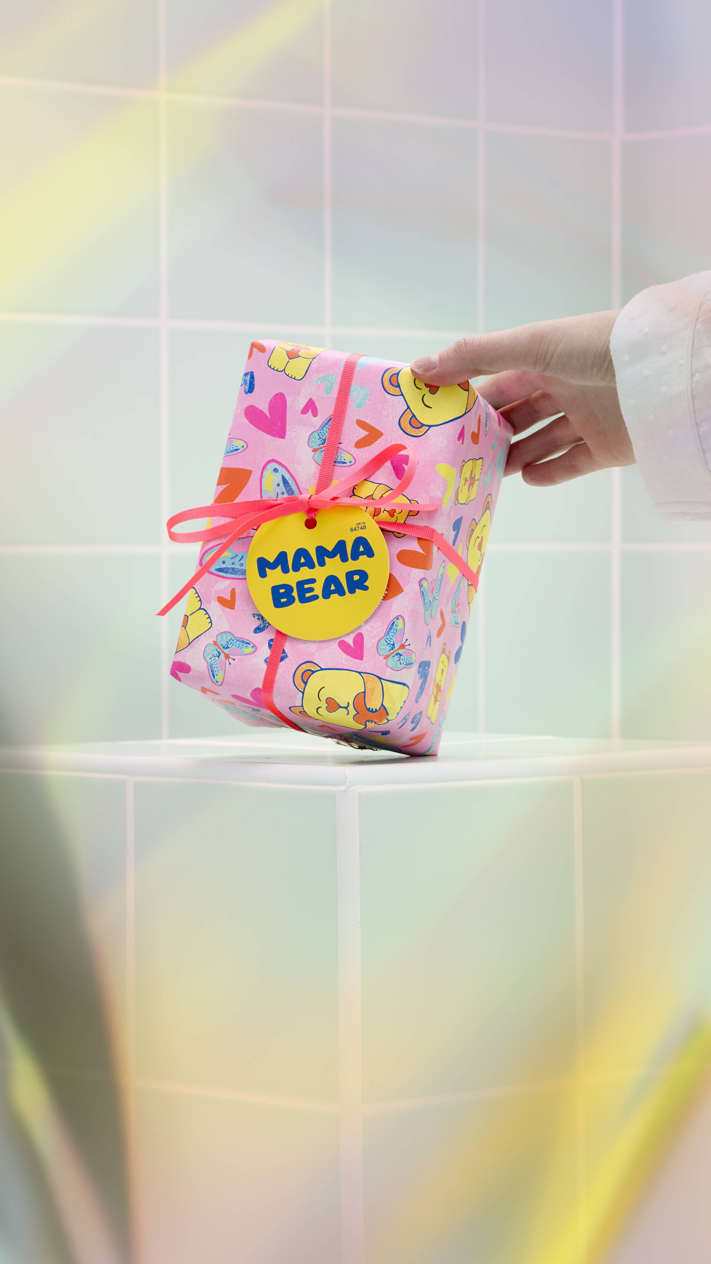 A model's hand reaches out to touch a Lush "Mama Bear" gift, which is wrapped in pink paper decorated with colourful bears, butterflies, and hearts. The gift is tied with a pink ribbon and a yellow tag against a background of light green tiles.