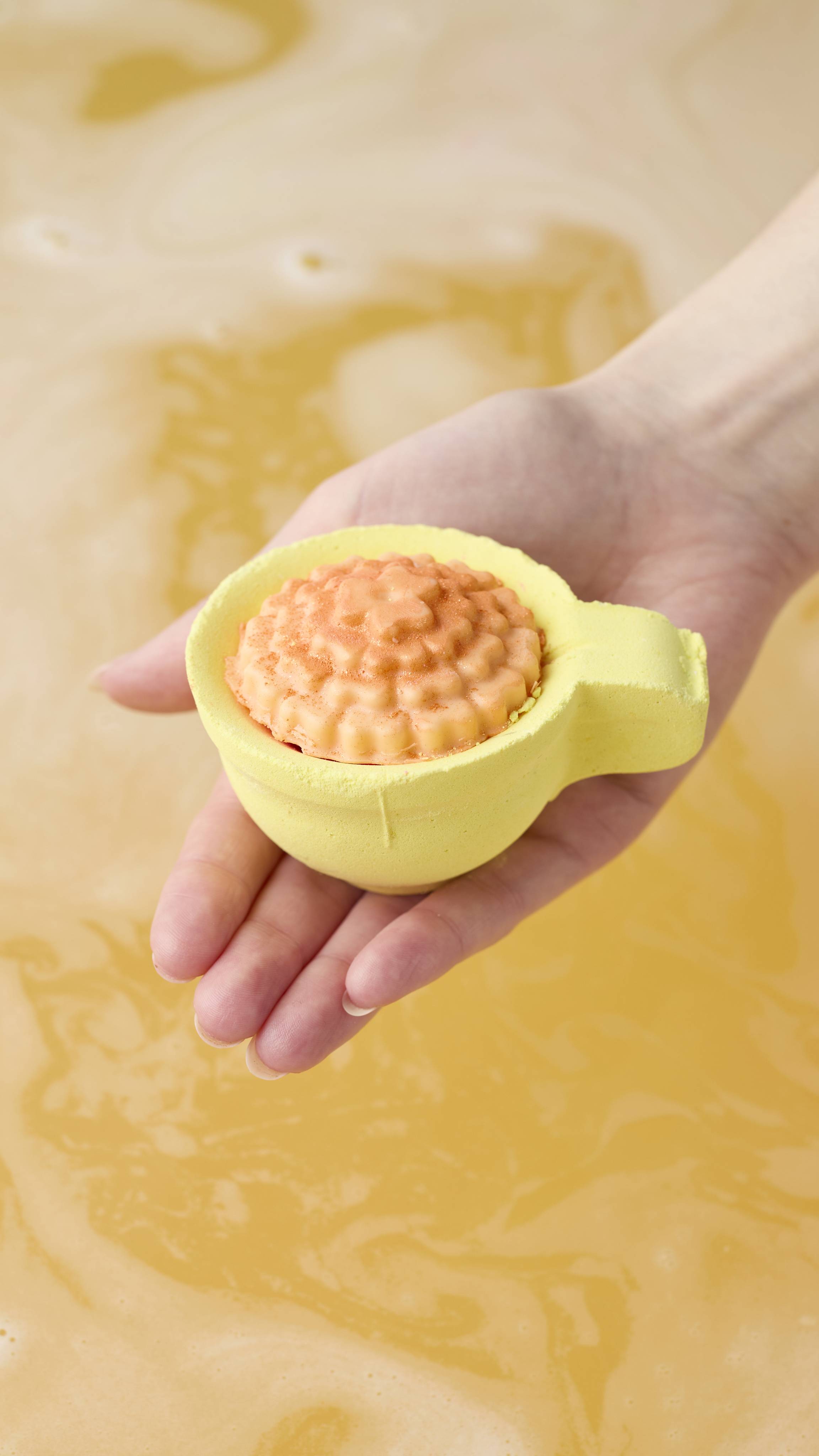 A hand holds the yellow teacup-shaped bath bomb, filled with a textured orange center, above warm golden bathwater. The water below swirls with matching yellow hues, suggesting a tea-infused bath.