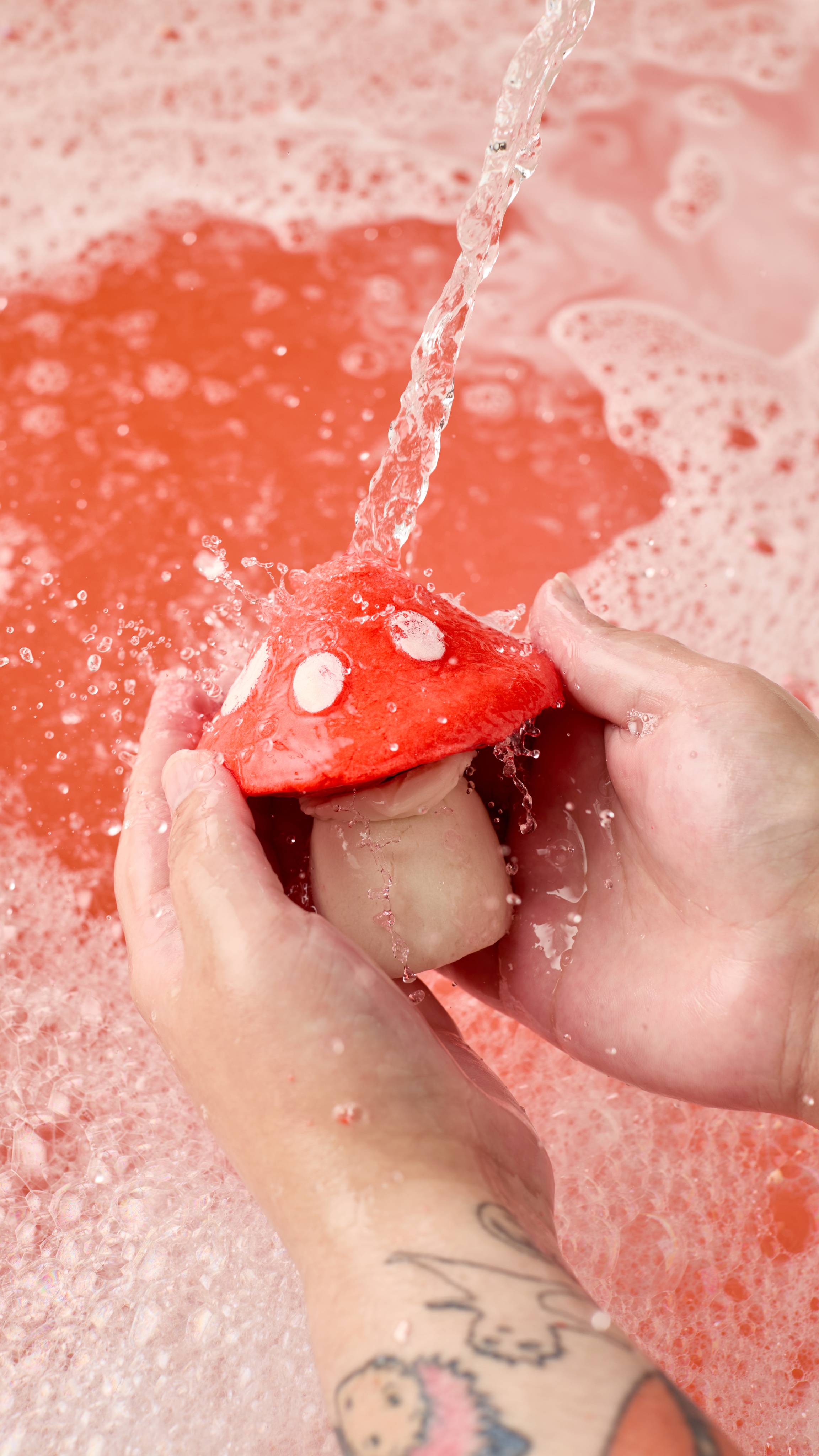 The image shows two light-toned hands holding a red toadstool-shaped 'Midnight Magic' bubble bar under running water. A stream pours onto the bar, generating abundant bubbles over red bath water.