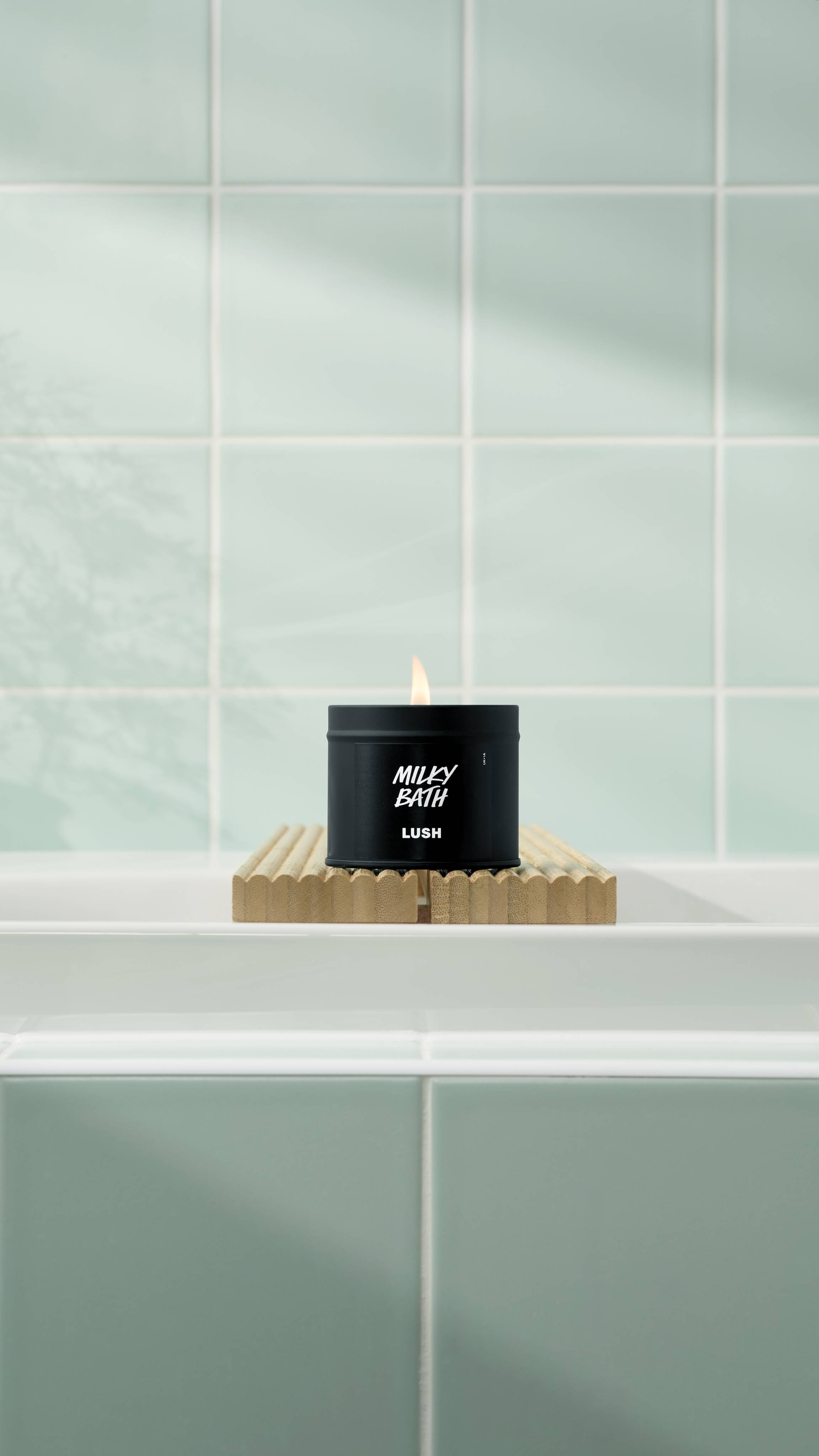 A lit Milky Bath candle in a matte black tin sits on a fluted wooden bath bridge over a white tub. The background features soft mint green square tiles with a gentle shadow of a plant. 