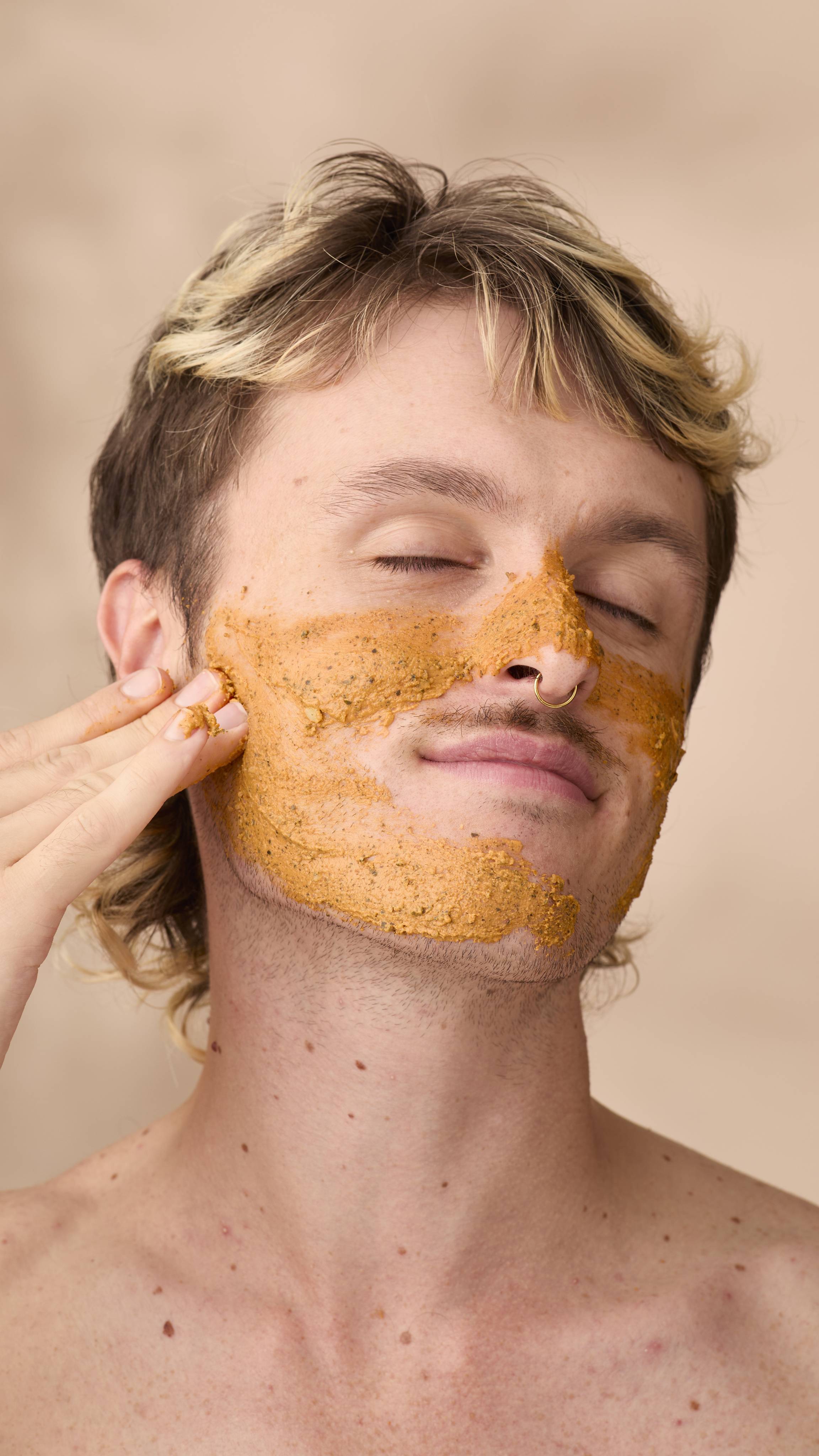 A close-up of a person with blonde-highlighted hair and eyes closed, smiling gently while applying an orange-brown Lush Mr Pumpkin Face mask to their jawline. A textured layer of the mask covers their cheeks and nose. Beige background.