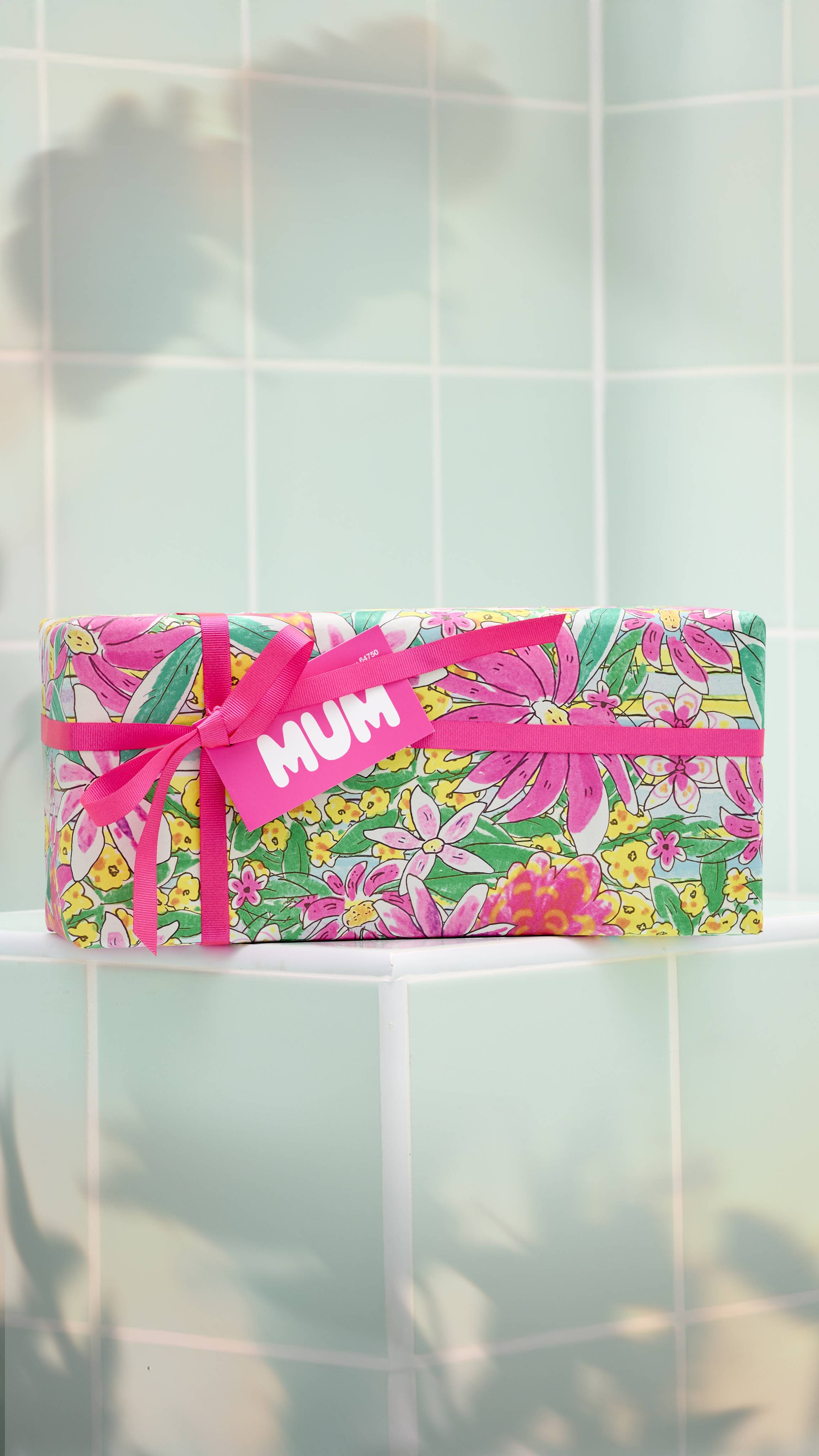 The image shows a rectangular gift wrapped in bright, floral-patterned paper with pink, yellow, and blue flowers. It is tied with a vibrant pink ribbon and features a matching pink "MUM" tag, set against a background of pale green bathroom tiles.
