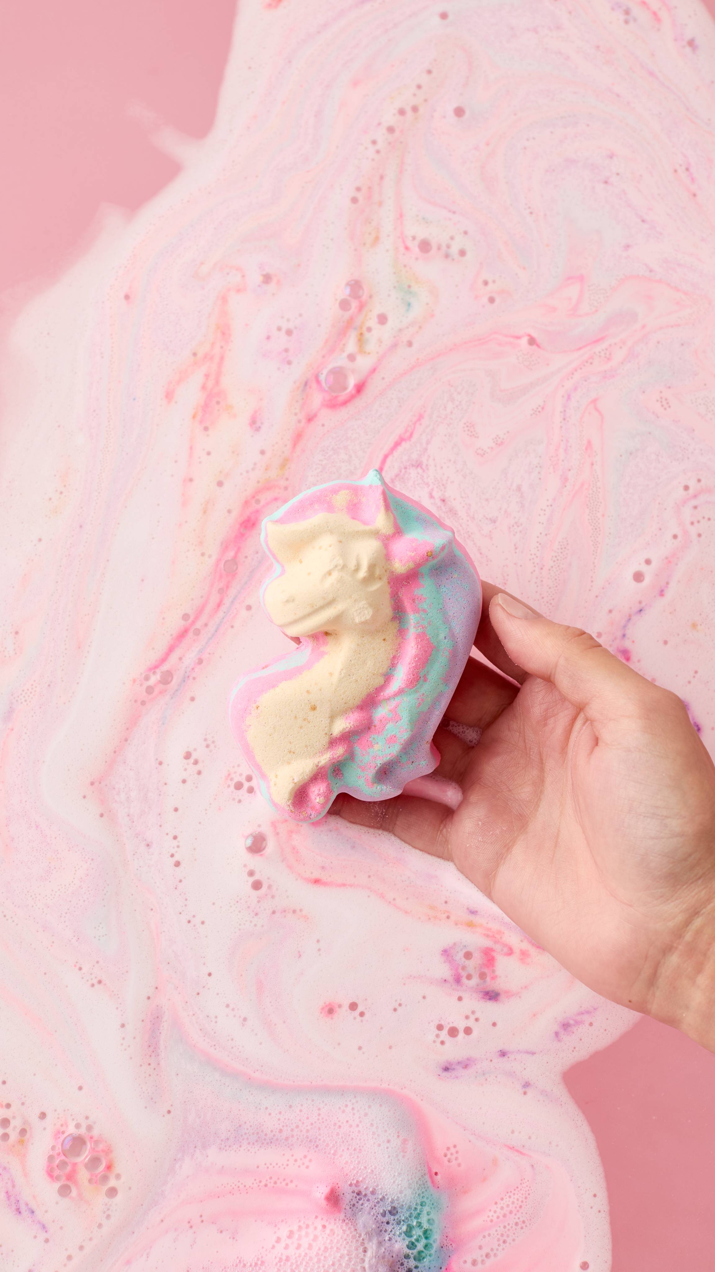 The image shows a light-toned hand cupping the My Little Unicorn bath bomb with a pink and blue mane. The product is held just above a vibrant pink bath with foamy bubbles and swirls.