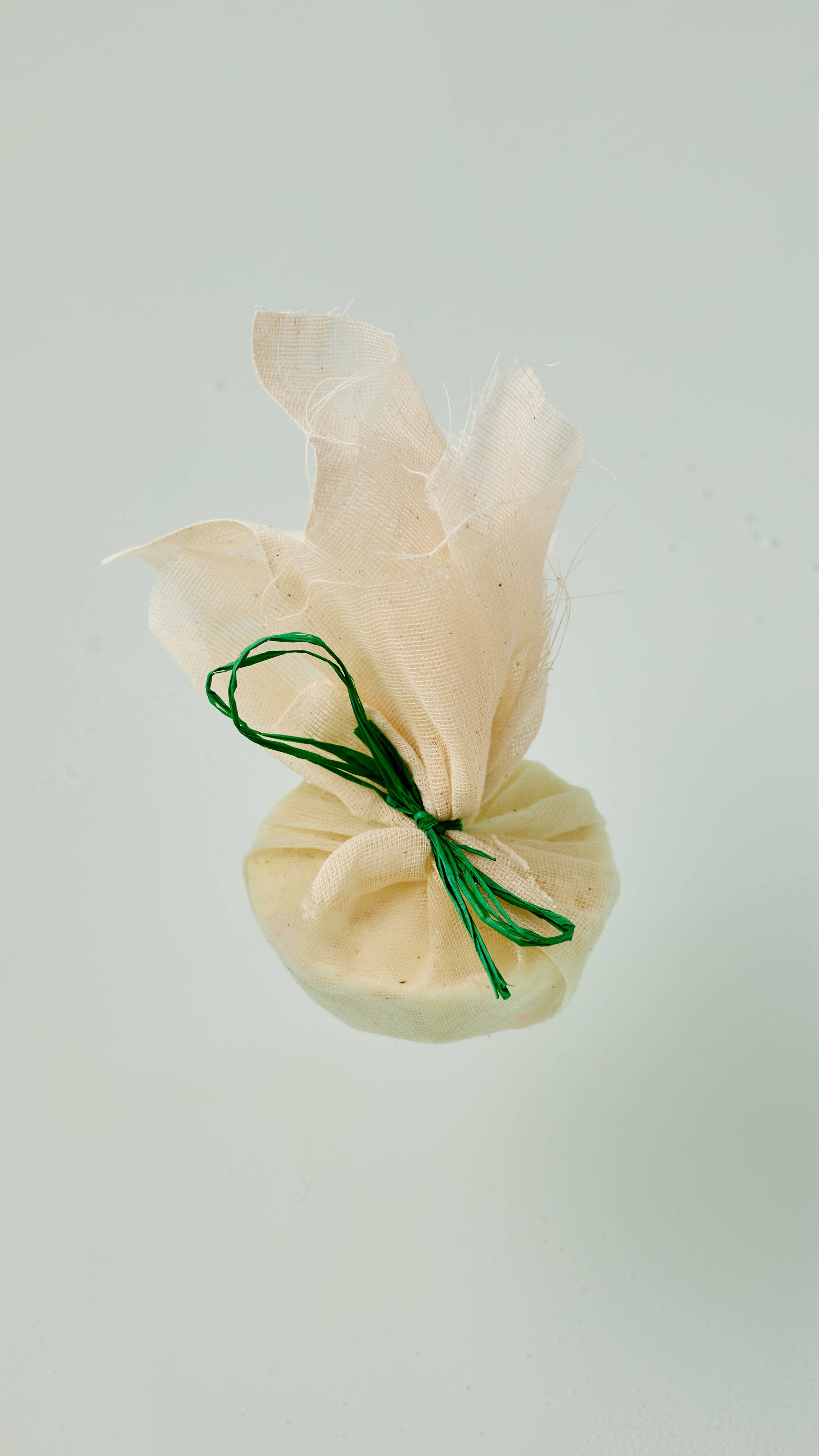The image shows the Narcotick solid bath oil. A roughly textured muslin fabric pouch, gathered at the middle and tied with a vibrant green string. The top of the fabric flares out above the rounded product, which rests on clear water.