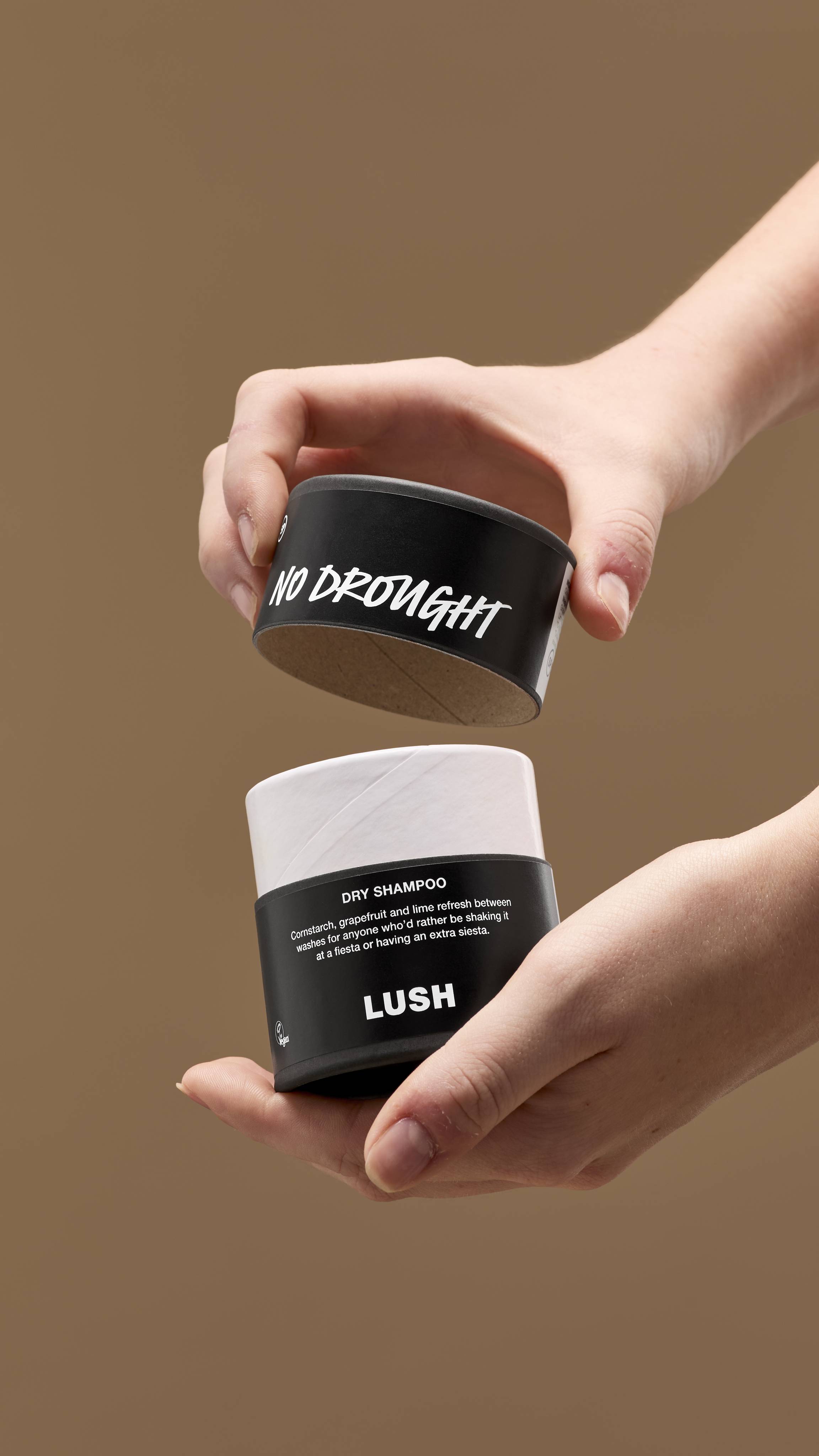 The image shows a pair of hands opening a black and white cardboard tub of 'No Drought' dry shampoo. The lid has white handwritten text, with the tub's black label reading 'DRY SHAMPOO' and 'LUSH', against a solid beige background.
