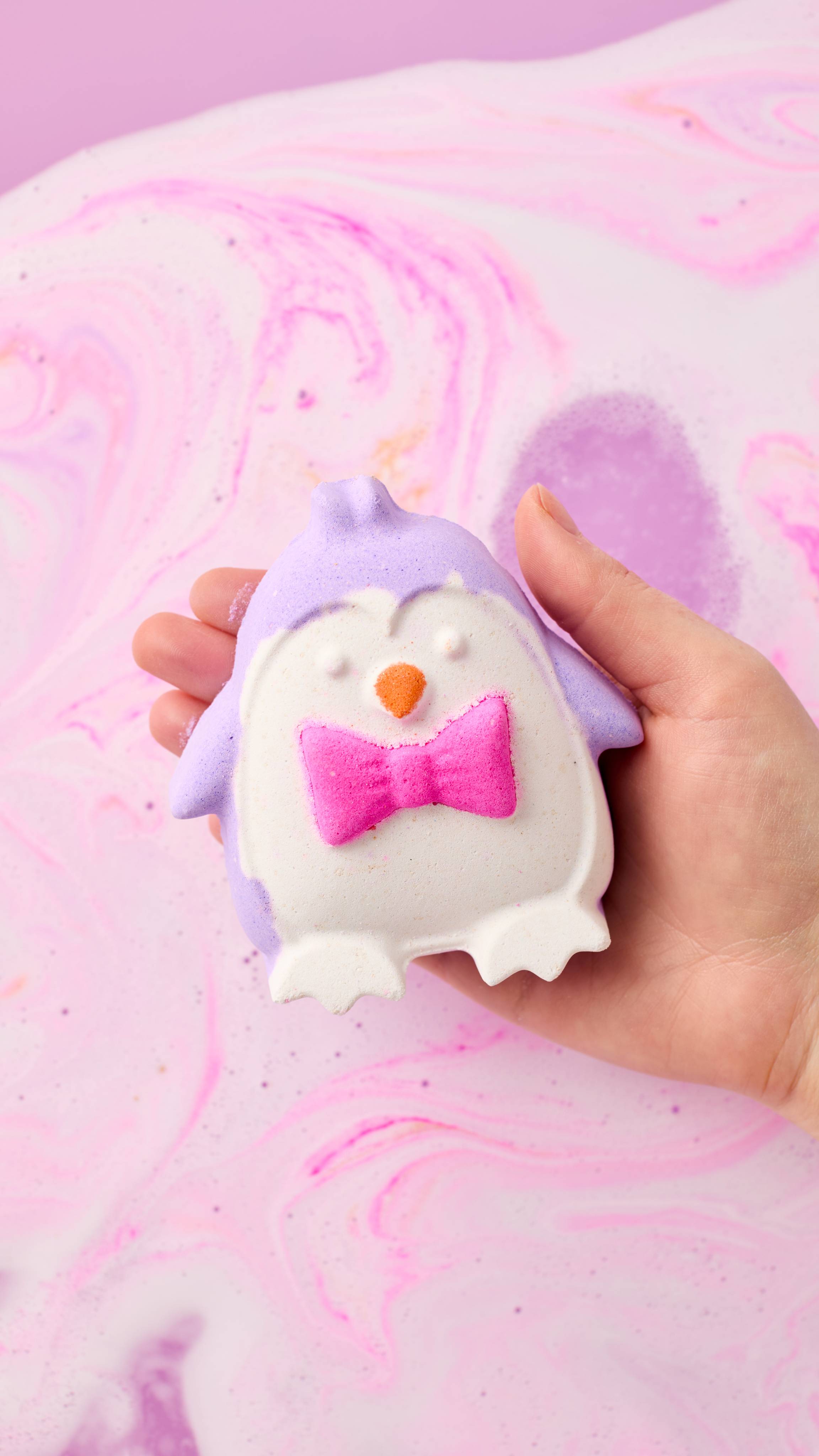 A hand holds a lilac and white penguin-shaped bath bomb featuring a bright pink bow tie and orange beak over swirling pastel pink and purple bathwater.