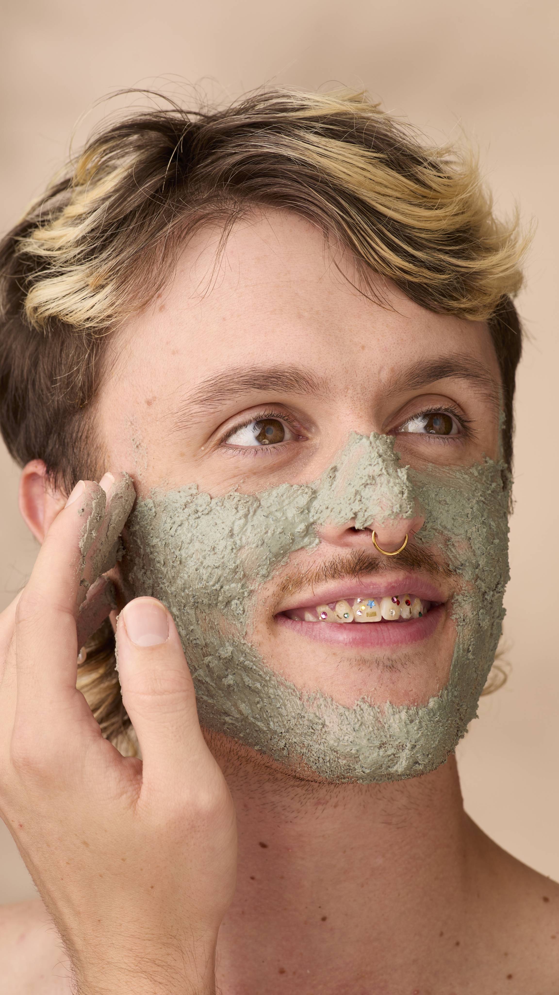 A person with blonde-highlighted hair smiles while applying a thick layer of greyish-green Persephone's Pomegranate face mask to their cheek. They wear a gold septum ring and have colourful gems on their teeth. Beige background.