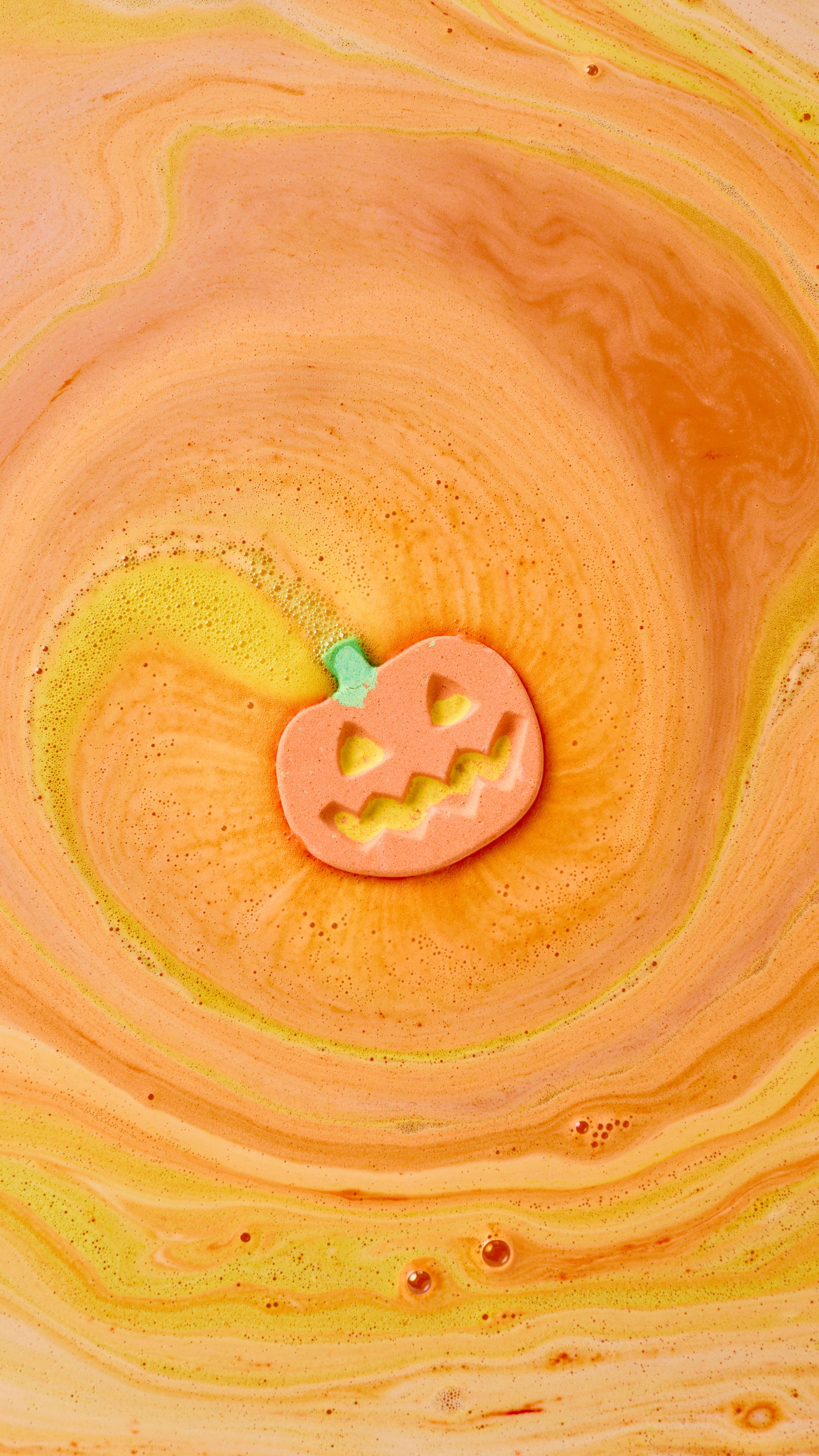 The image shows an orange 'Punkin Pumpkin' bath bomb, with a green stem and carved features, fizzing in swirling yellow and orange bath water. The water forms a vibrant spiral around the dissolving bath bomb.