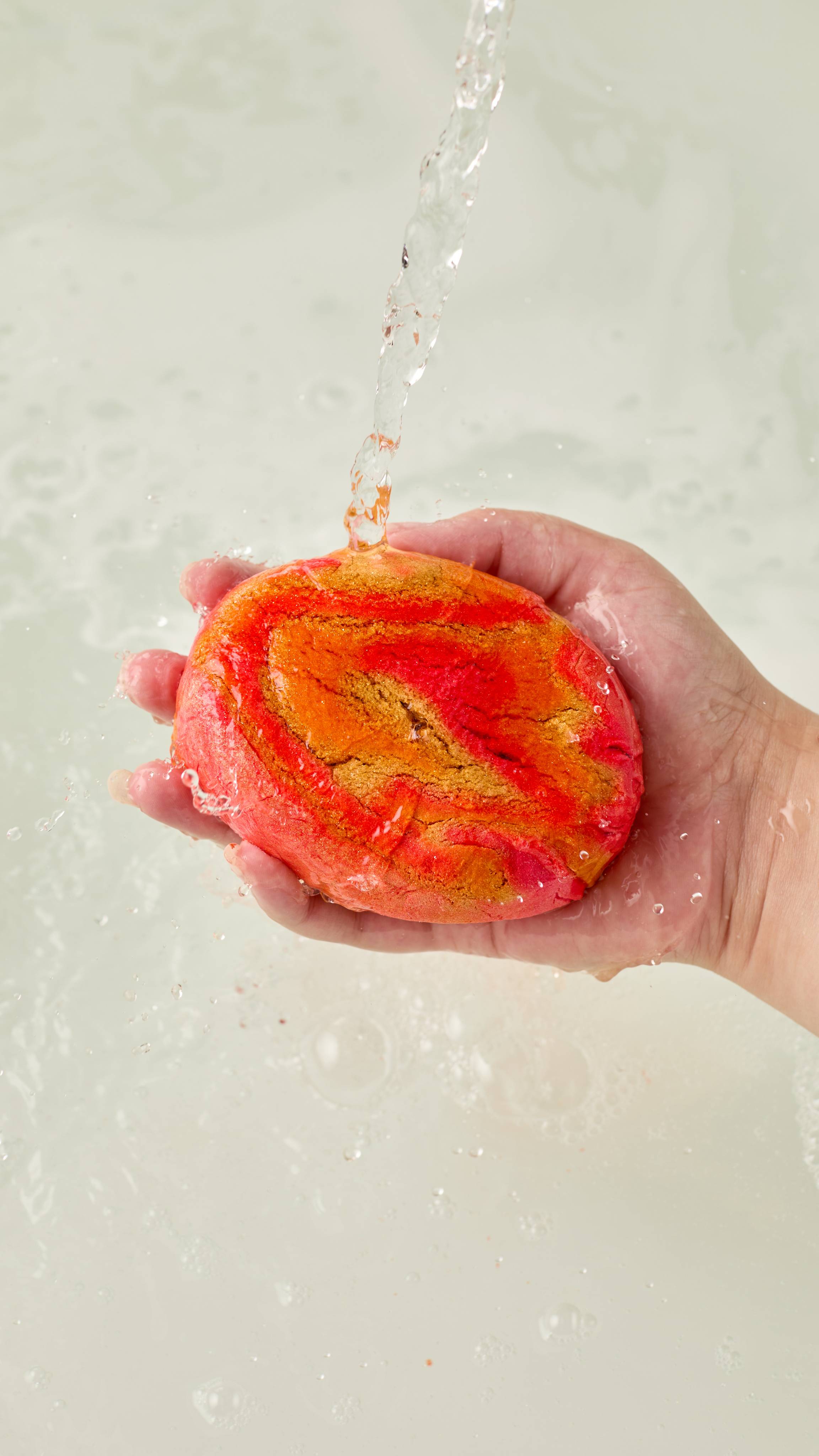 The image shows a light-toned hand holding a vibrant gold and orange swirled 'Pumpkin Spice' bubble bar. Water pours onto the bar, generating abundant white bubbles and thick foam across the bath.