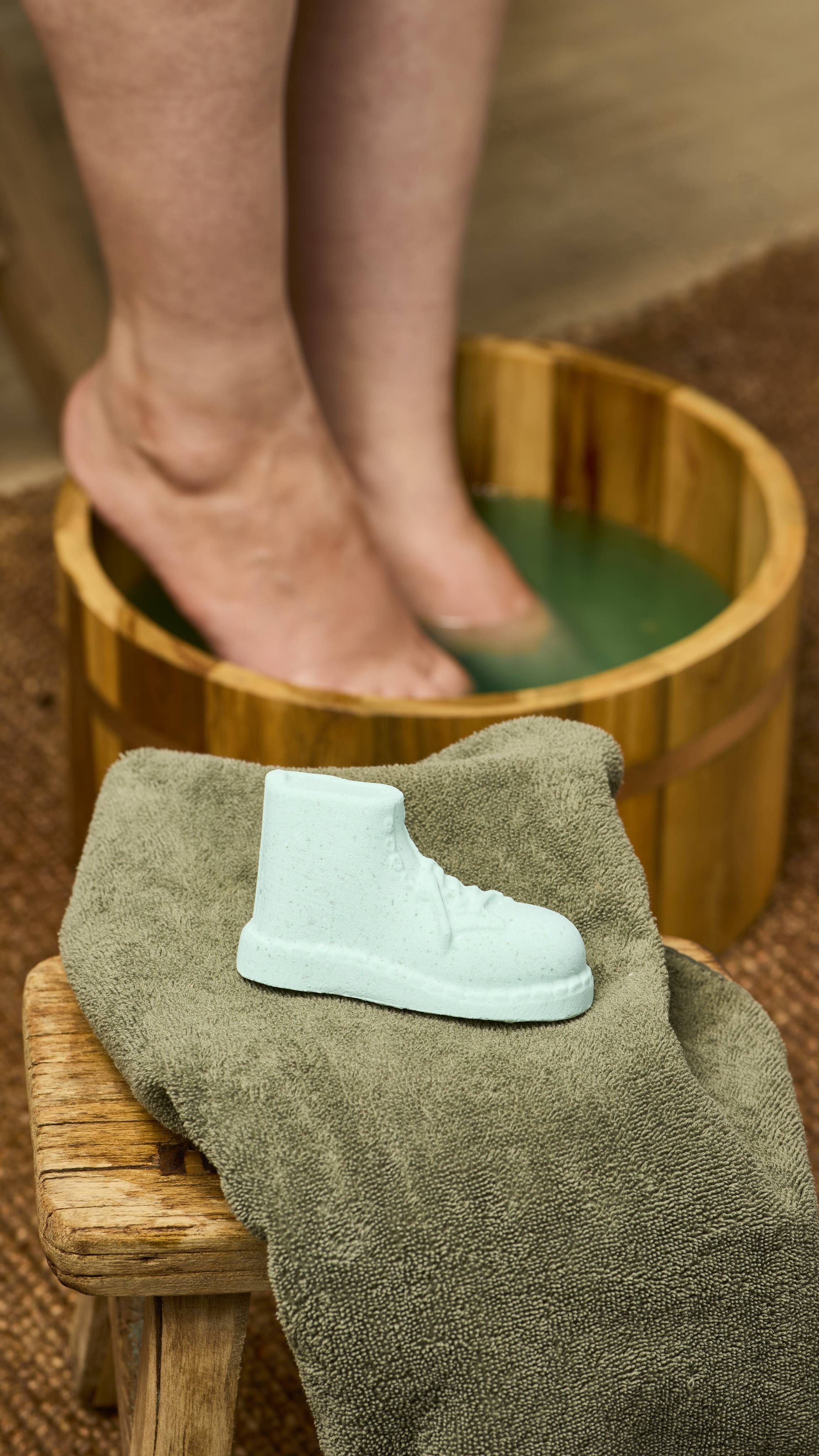 The image shows the Put The Boot In foot soak product resting on a folded, khaki-coloured towel. In the blurred background, feet are soaking in a wooden tub filled with green water, on a woven mat.