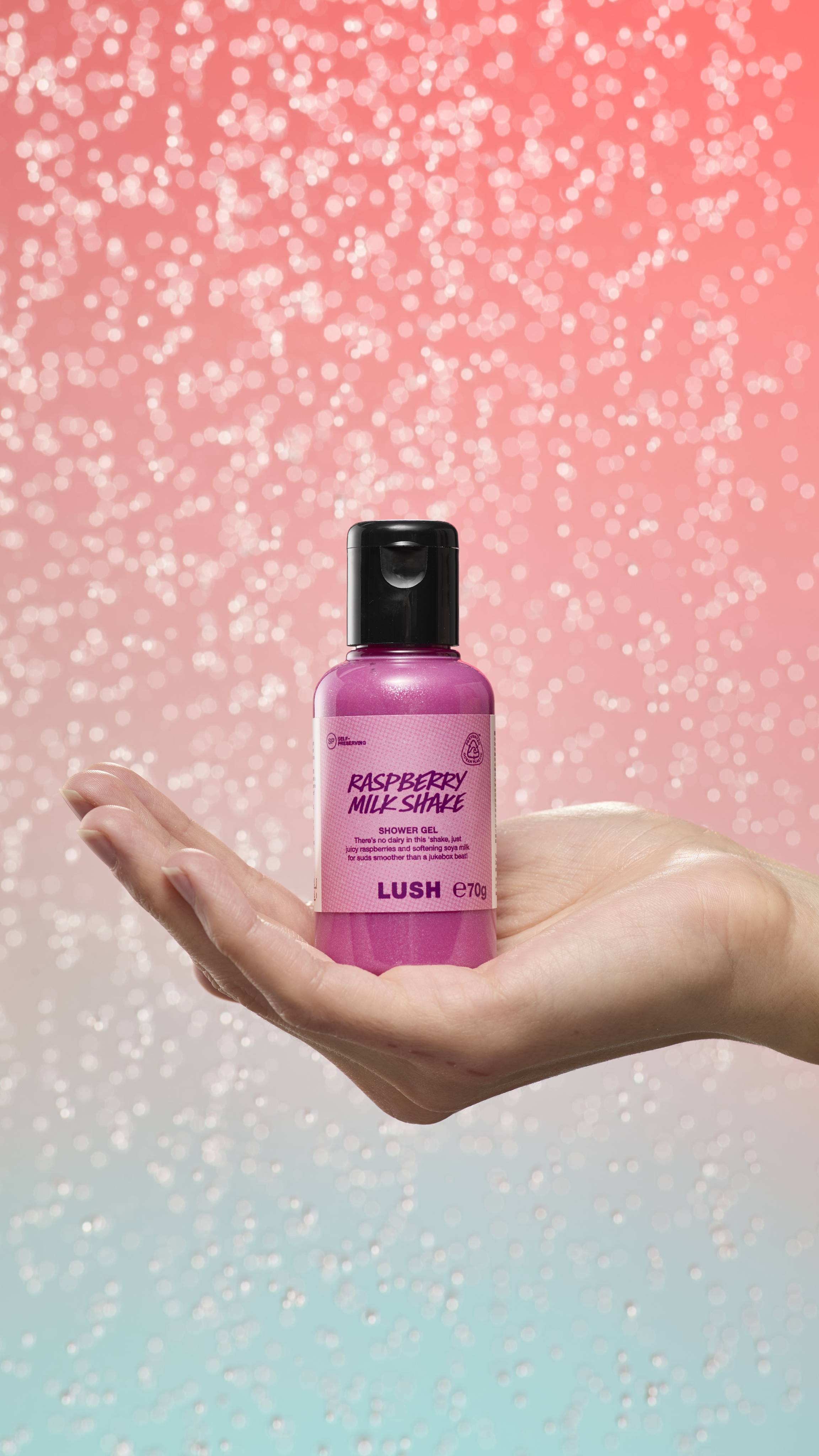 Hand holds a small pink Raspberry Milkshake shower gel bottle against a shimmering pink-peach gradient. The label is simple with dark pink text and the LUSH logo.