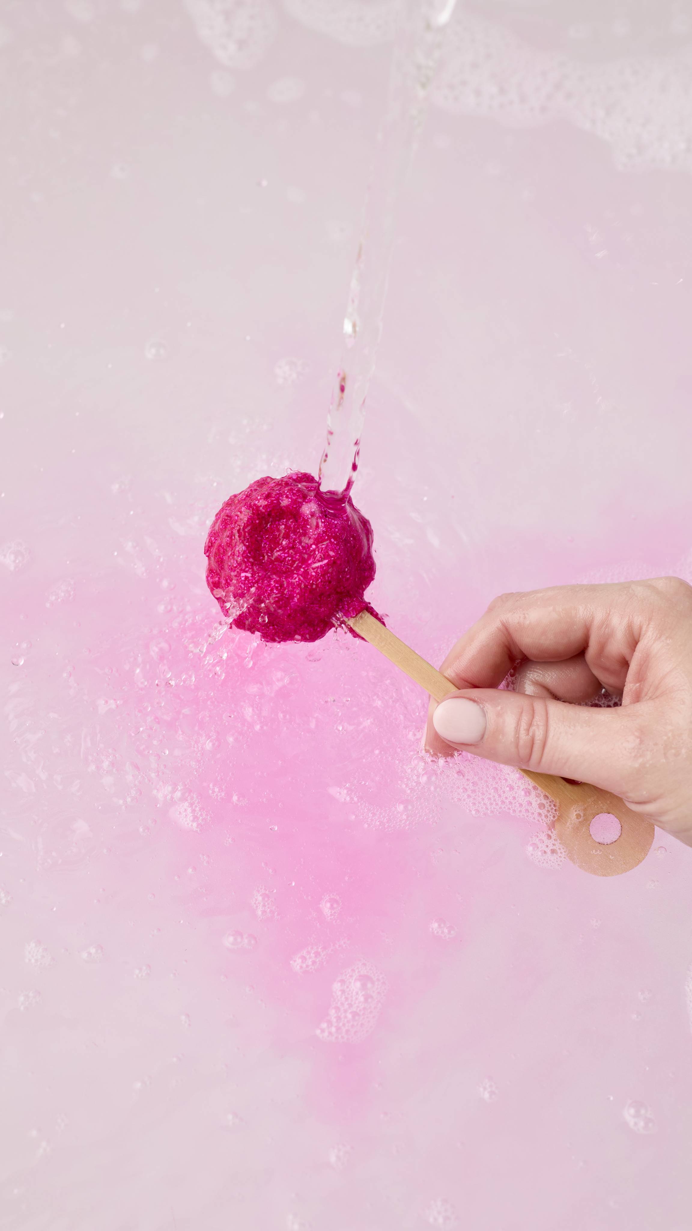 A hand holds a Raspberry mini bath wand under a stream of running water. The water hits the shimmering magenta base, sending a vibrant pink swirl into the pale pink, foamy bath water below.
