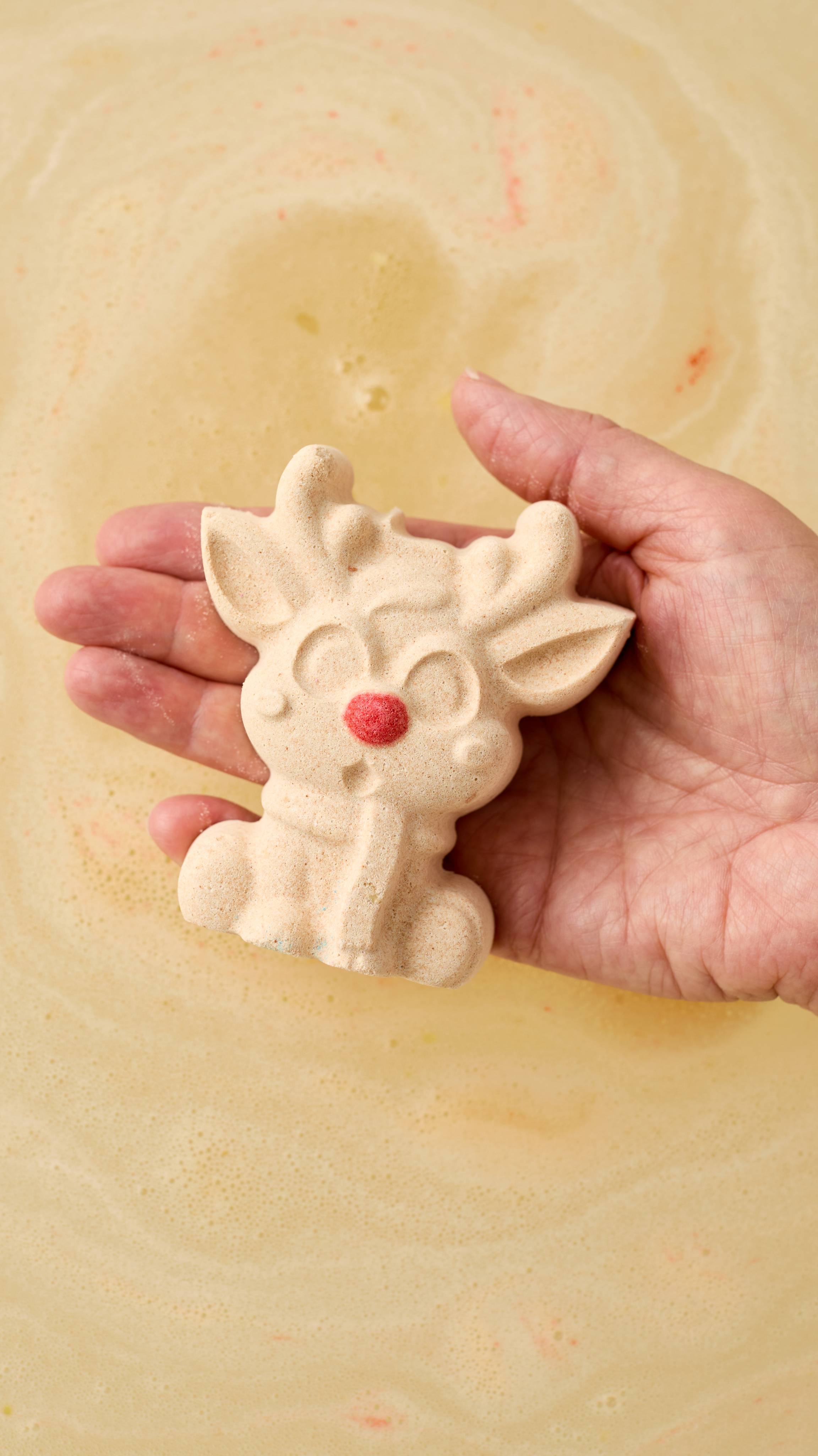 The image shows a light-toned hand holding a pale cream, speckled Reindeer bath bomb with a red nose. The hand is cupped over swirling yellow bath water, with a light foam on the surface.