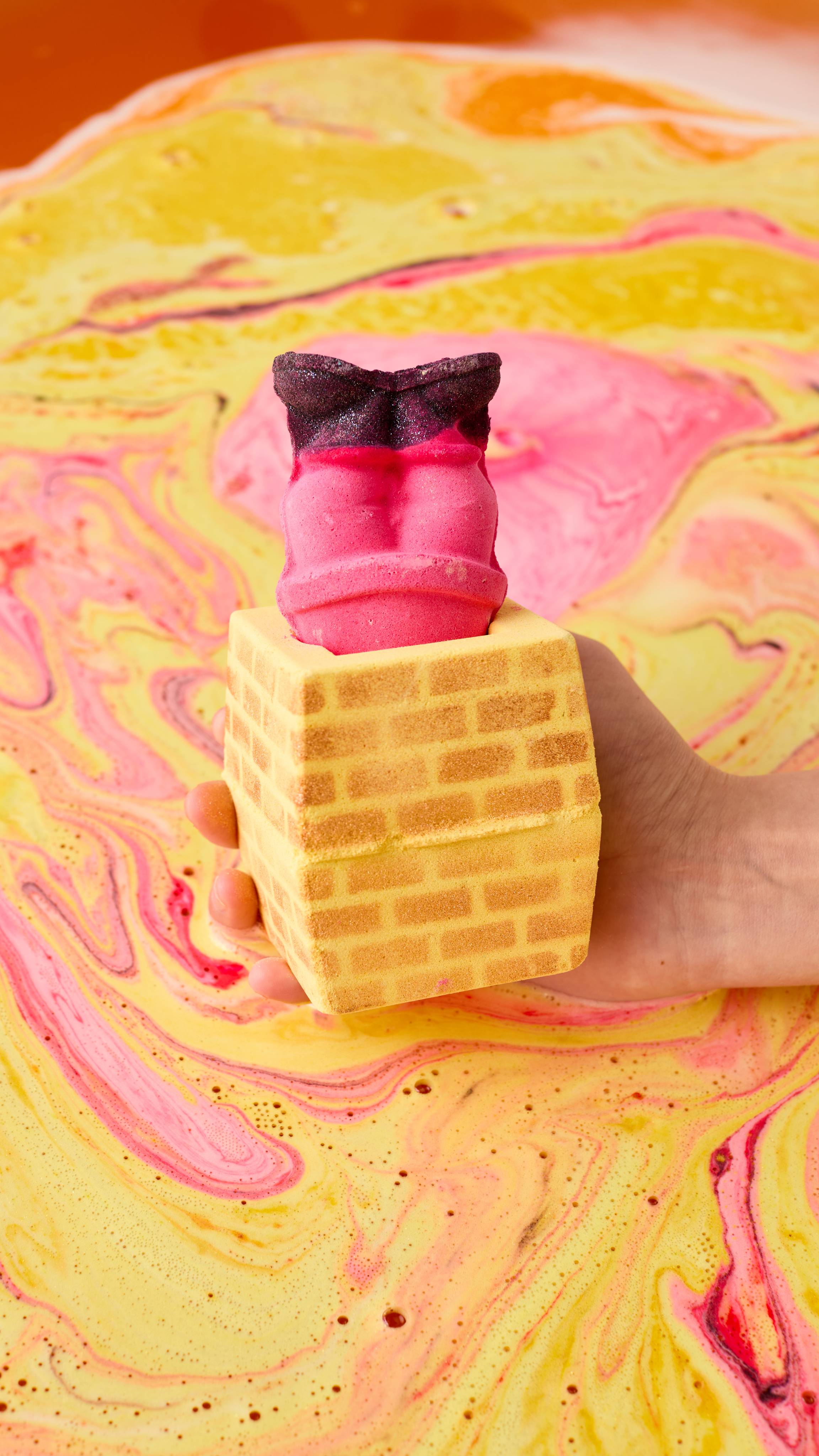 The image shows a hand holding the Stuck In The Chimney bath bomb over swirling bath water. The yellow brick chimney has red legs and shimmering black boots sticking out of the top, and the water is marbled with yellow and pink.