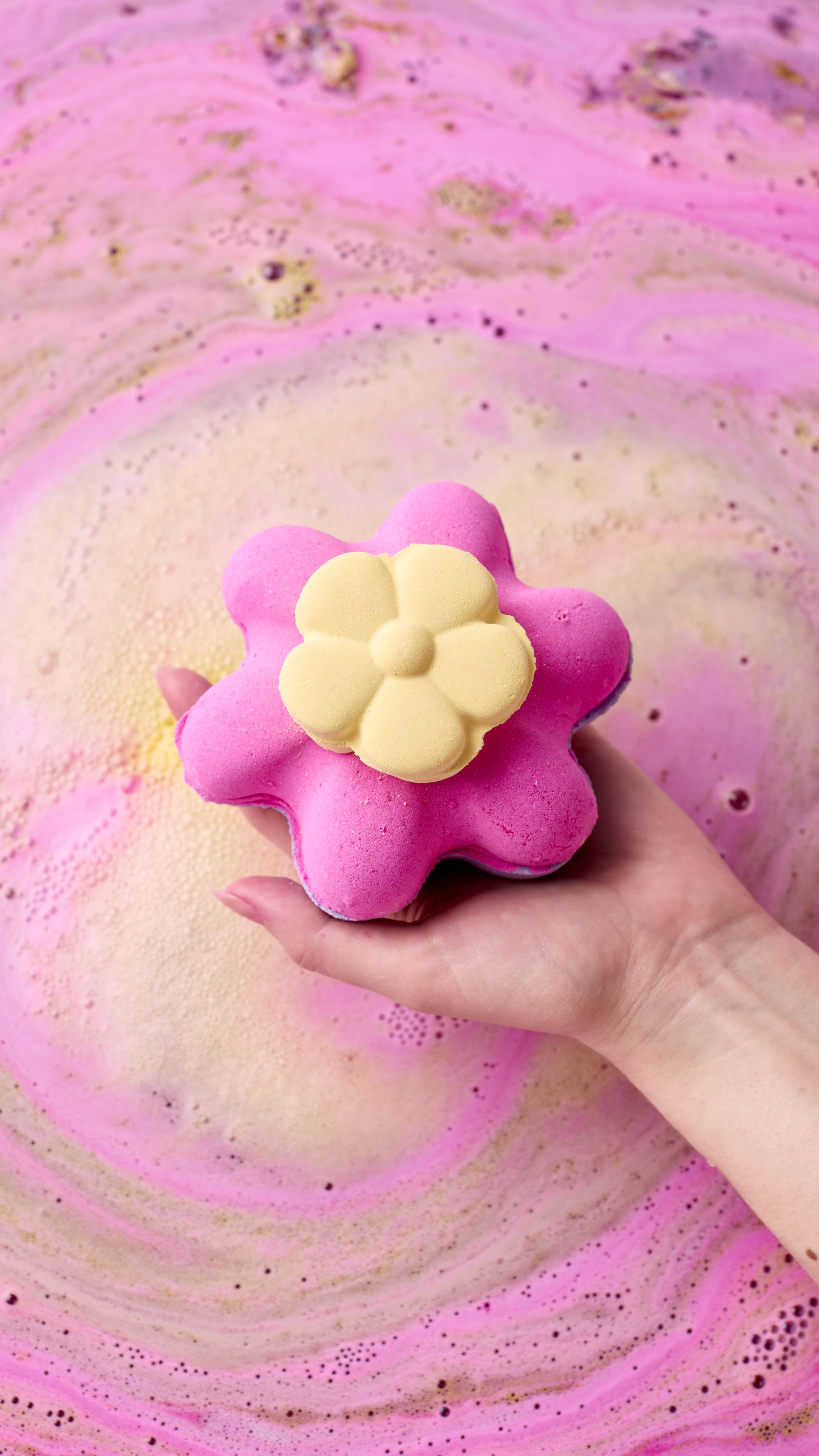 A hand holds the Secret Garden Bombshell bath bomb over swirling pink bathwater. The bath bomb is shaped like a vibrant magenta flower topped with a smaller yellow flower center.