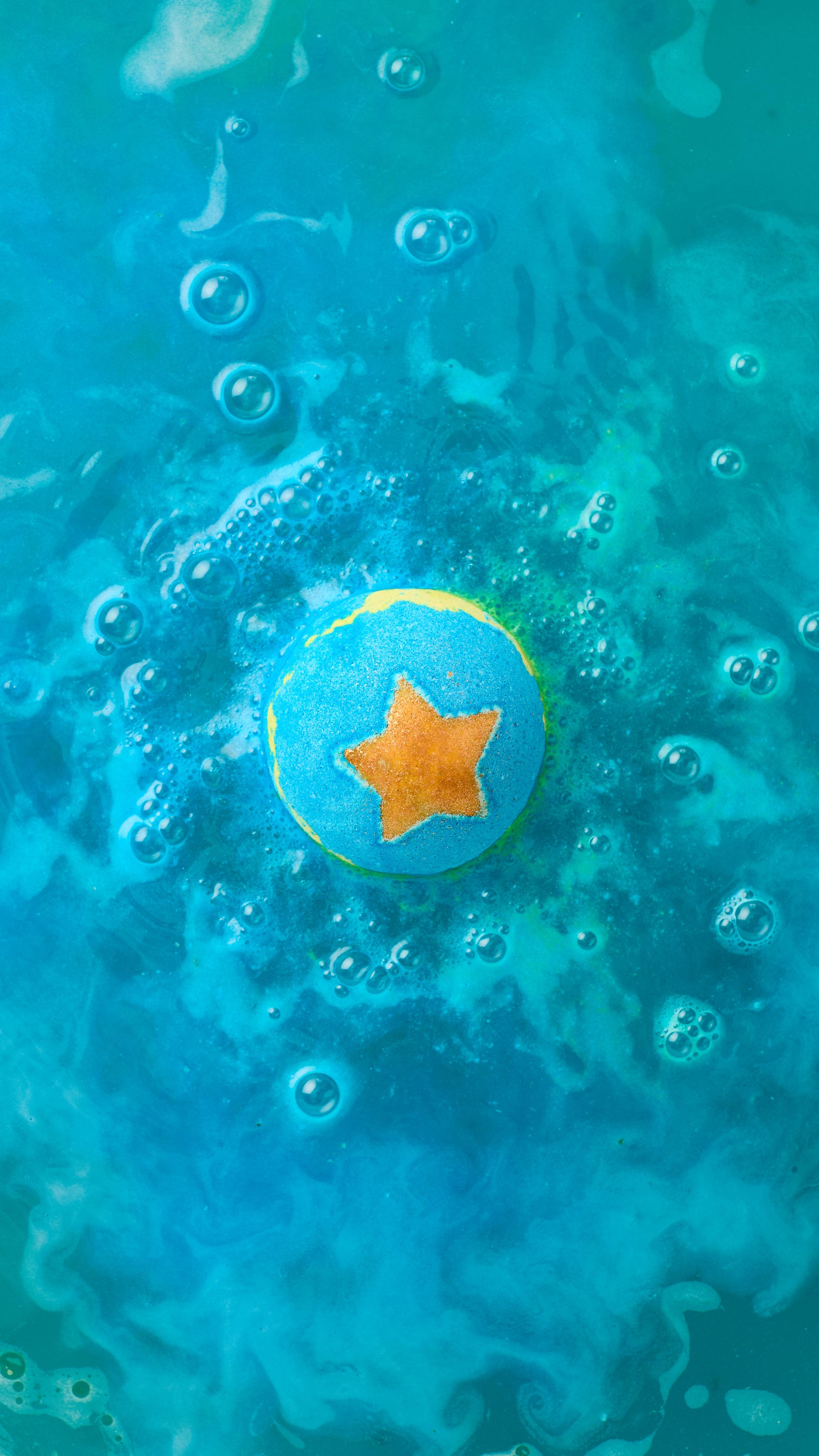 The image shows a blue, star-topped 'Shoot For The Stars' bath bomb fizzing in the water. Vibrant hues of dark and light blue swirl around the product, mixing with abundant foam and bubbles.