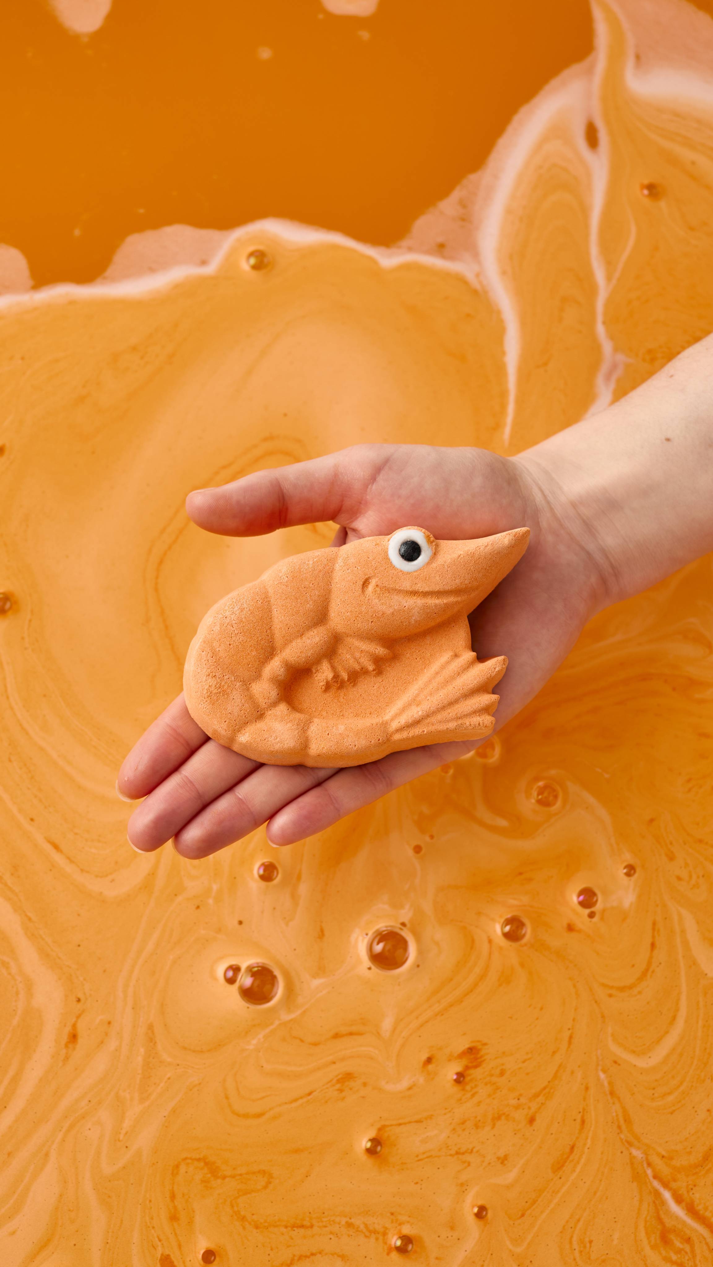 A model's hand holds a bright orange, shrimp-shaped bath bomb over a bathtub filled with deep orange water. The water’s surface is covered in swirls of creamy, pale orange foam and small clusters of bubbles.