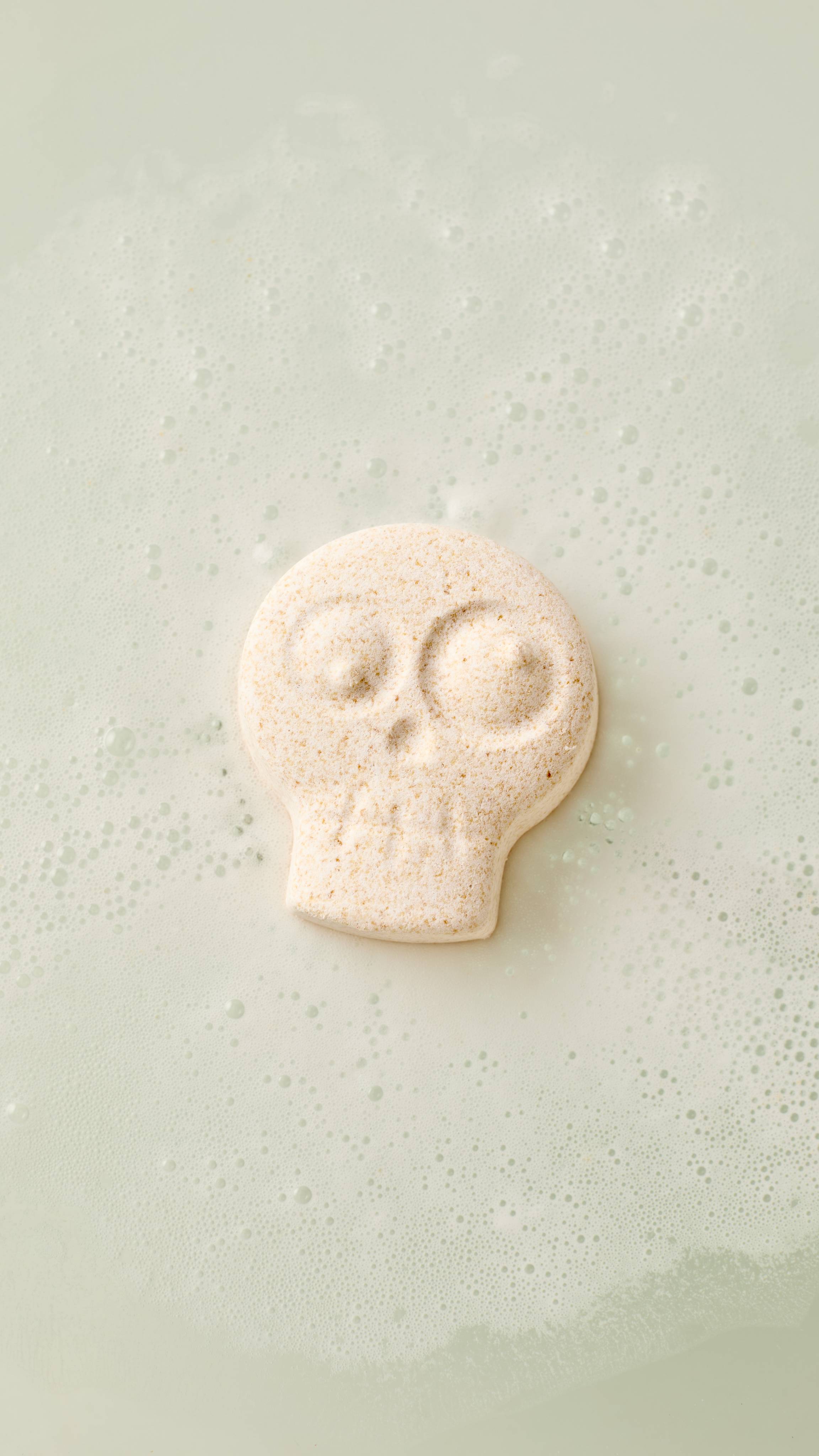 The image shows a pale, skull-shaped 'Skeleton - Monster Mash Head' bath bomb floating on the surface of creamy, bubbly bath water. The bomb features embossed eye sockets and nose details.