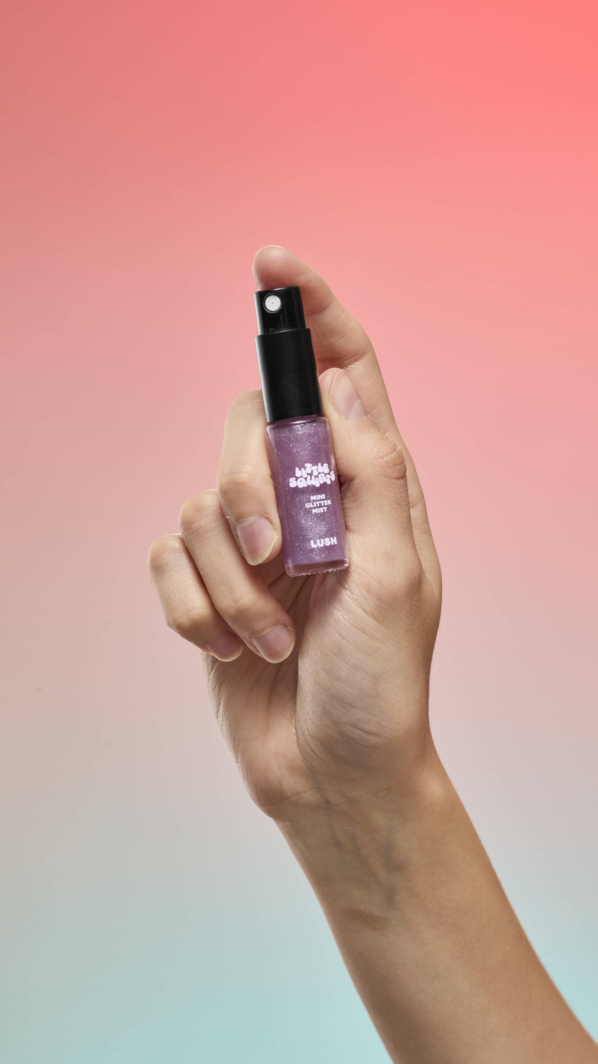 A hand holds a small bottle of purple Little Squirts Mini Glitter Mist against a peach-to-blue gradient background. The bottle features a black spray cap and a white LUSH logo.