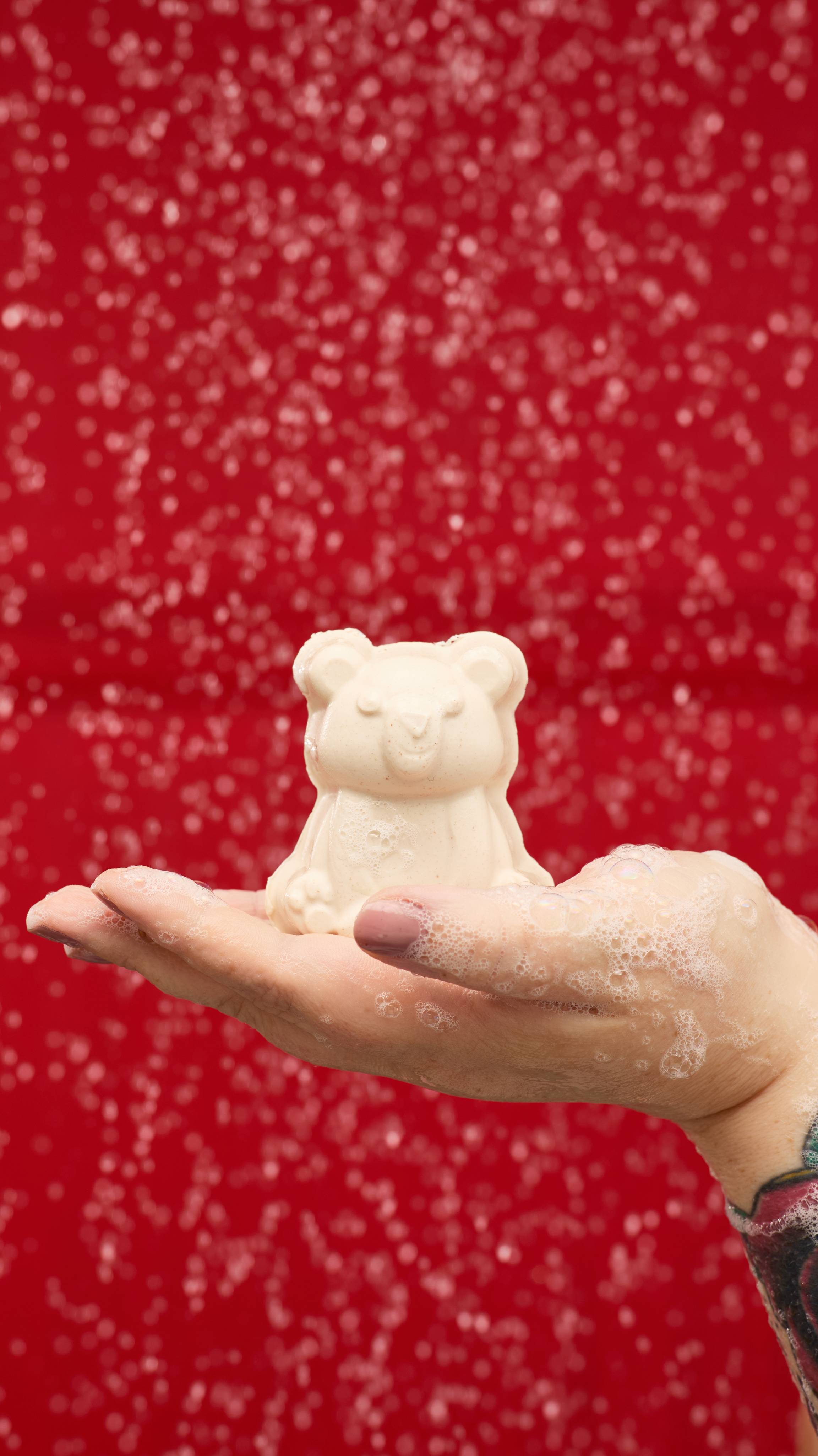 A person holds a white, Snow Bear soap in their palm, covered in a thick lather of suds. Water droplets and bubbles dot the hand, with a solid red background behind it.