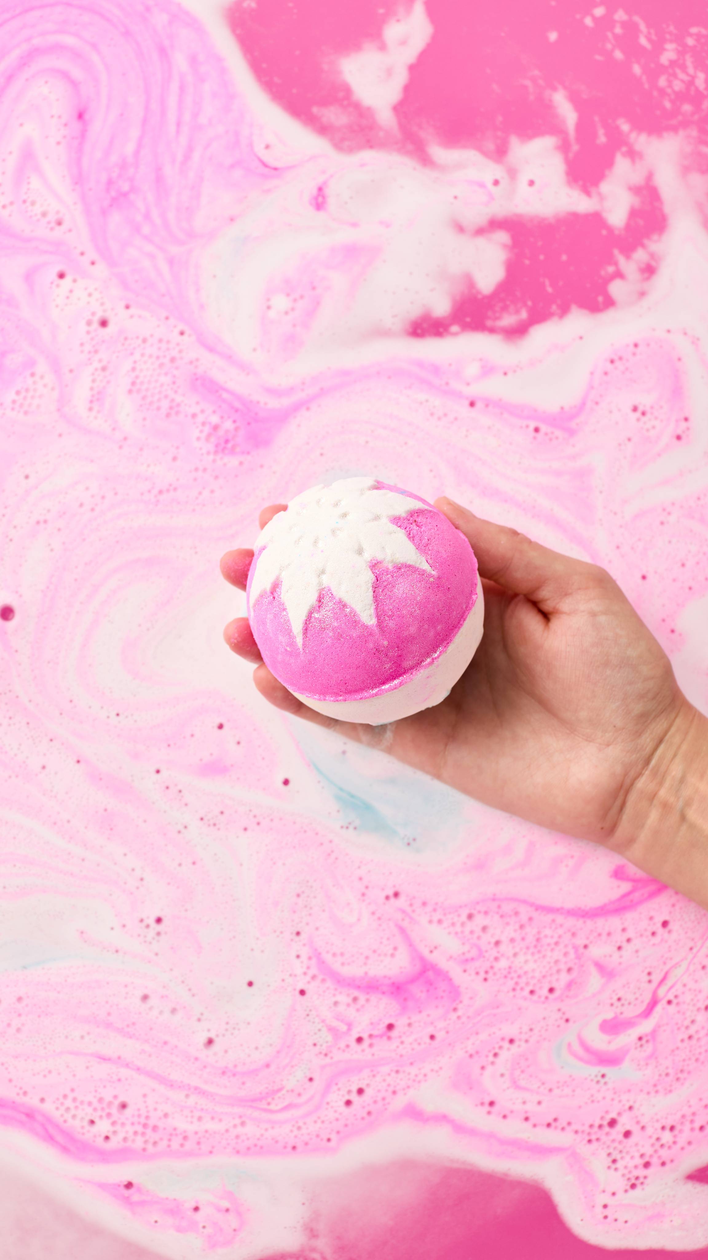 The image shows a hand cupping a pink and white spherical Snow Fairy bath bomb. The product, with its embossed snowflake, is held just above a vibrant pink bath with foamy bubbles and swirls.