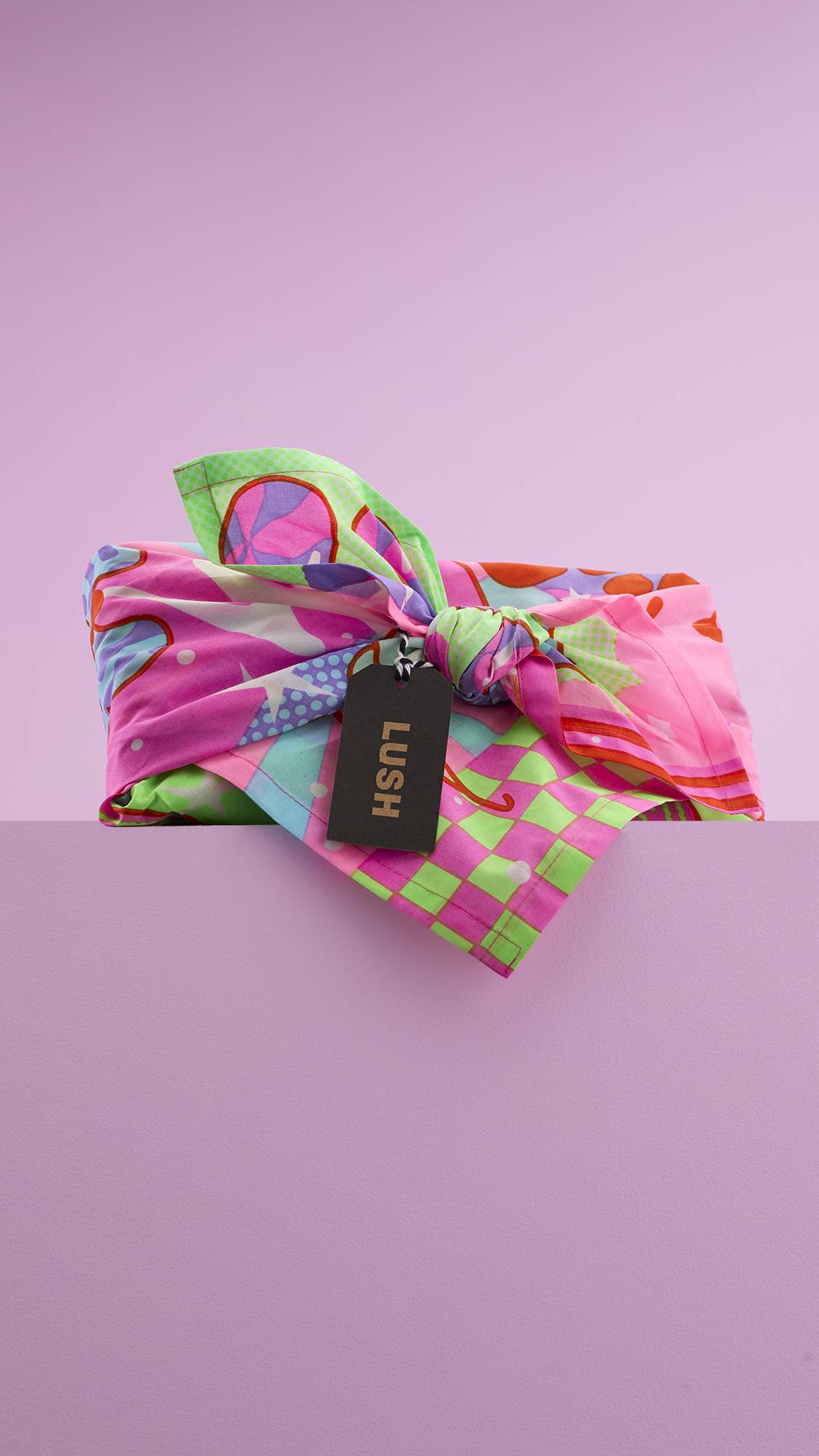 A vibrant gift wrapped in a Lush "Snow Fairy" Knot Wrap. The reusable fabric features a colorful collage of whimsical designs, tied with a knot and a black tag that reads "LUSH" in neon pink lettering.