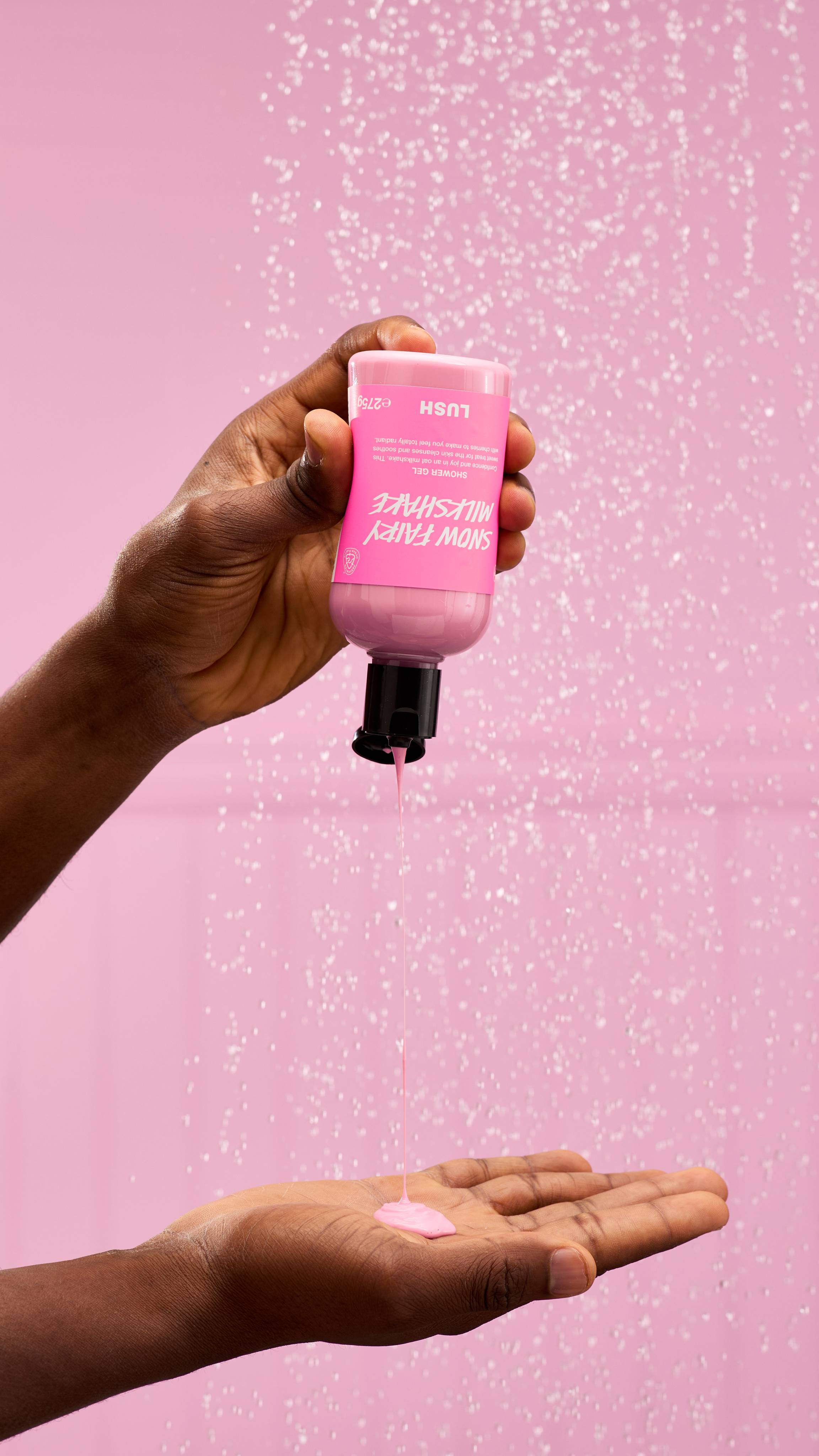 A warm-toned hand holds a bottle of pink Snow Fairy Milkshake shower gel. They pour the thick, pastel pink liquid from the bottle into their open palm. The background is a solid pink, and blurred shower droplets fall behind.