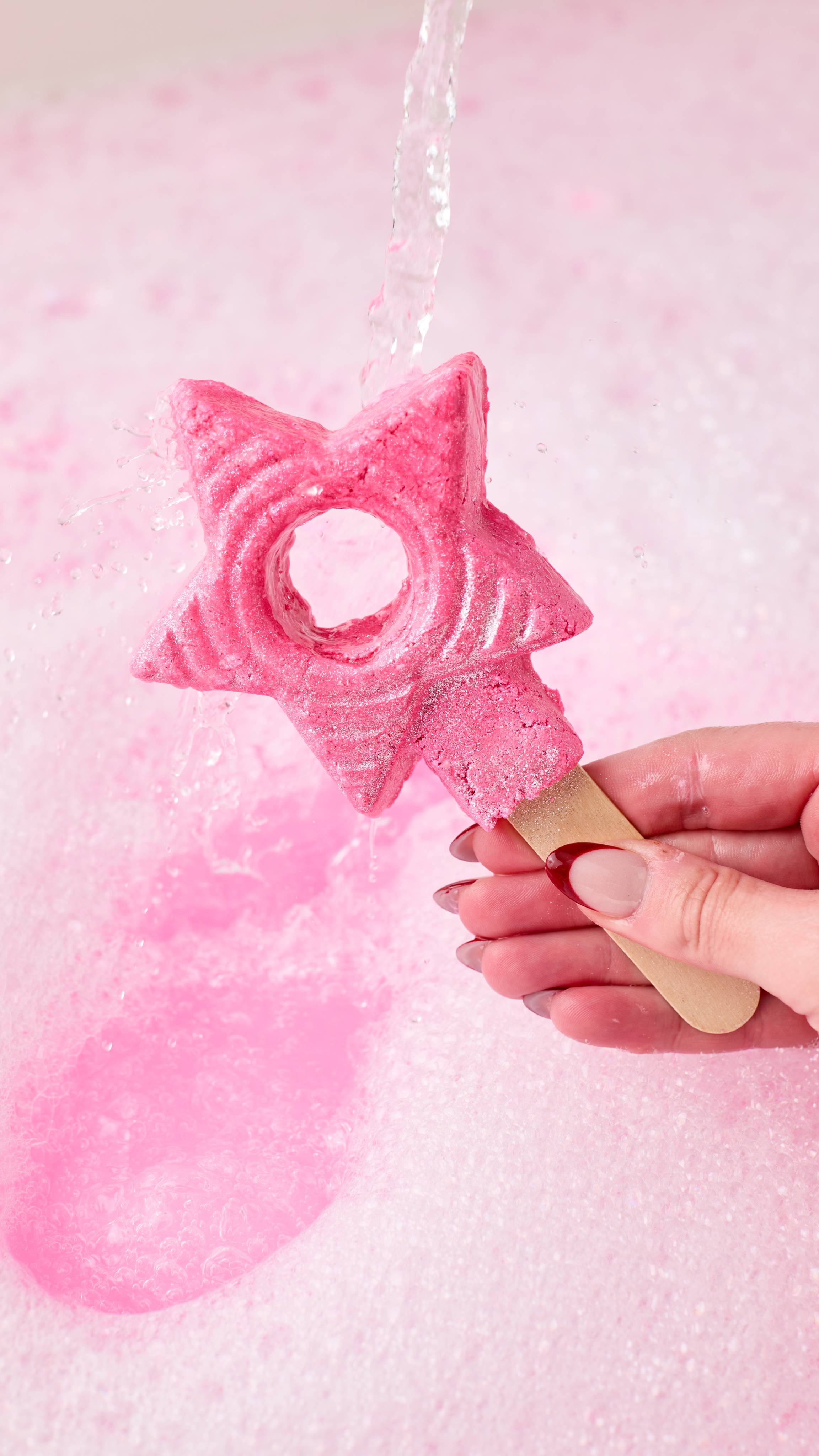 The image shows a hand holding the five-pointed star-shaped Snow Fairy Wand by the attached wooden stick. Water pours over the bubble wand and splashes off as it creates bubbles and pink water in the bath below.