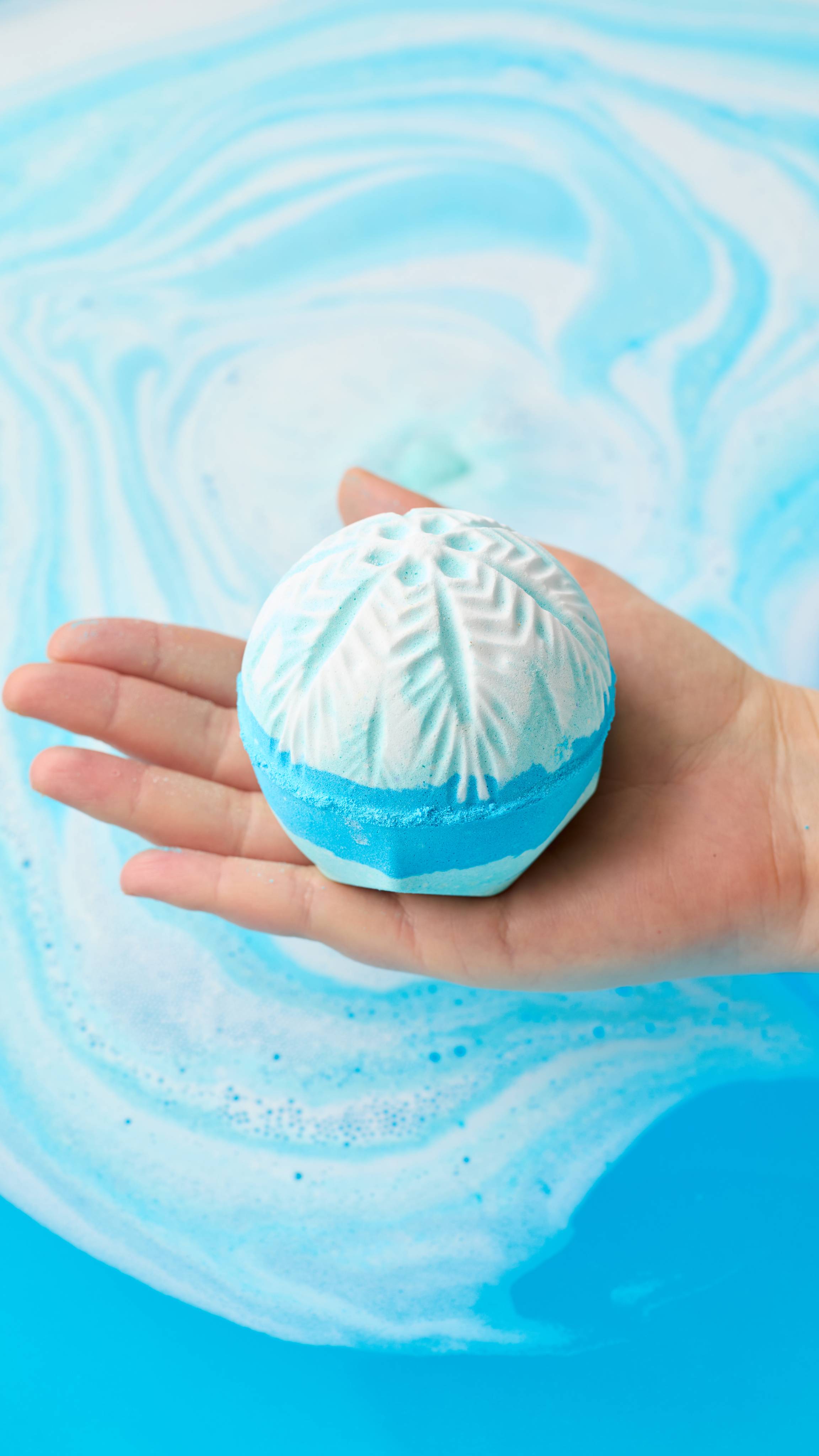 A hand holds a spherical white and light blue bath bomb embossed with a snowflake pattern over swirling, bright blue bathwater.